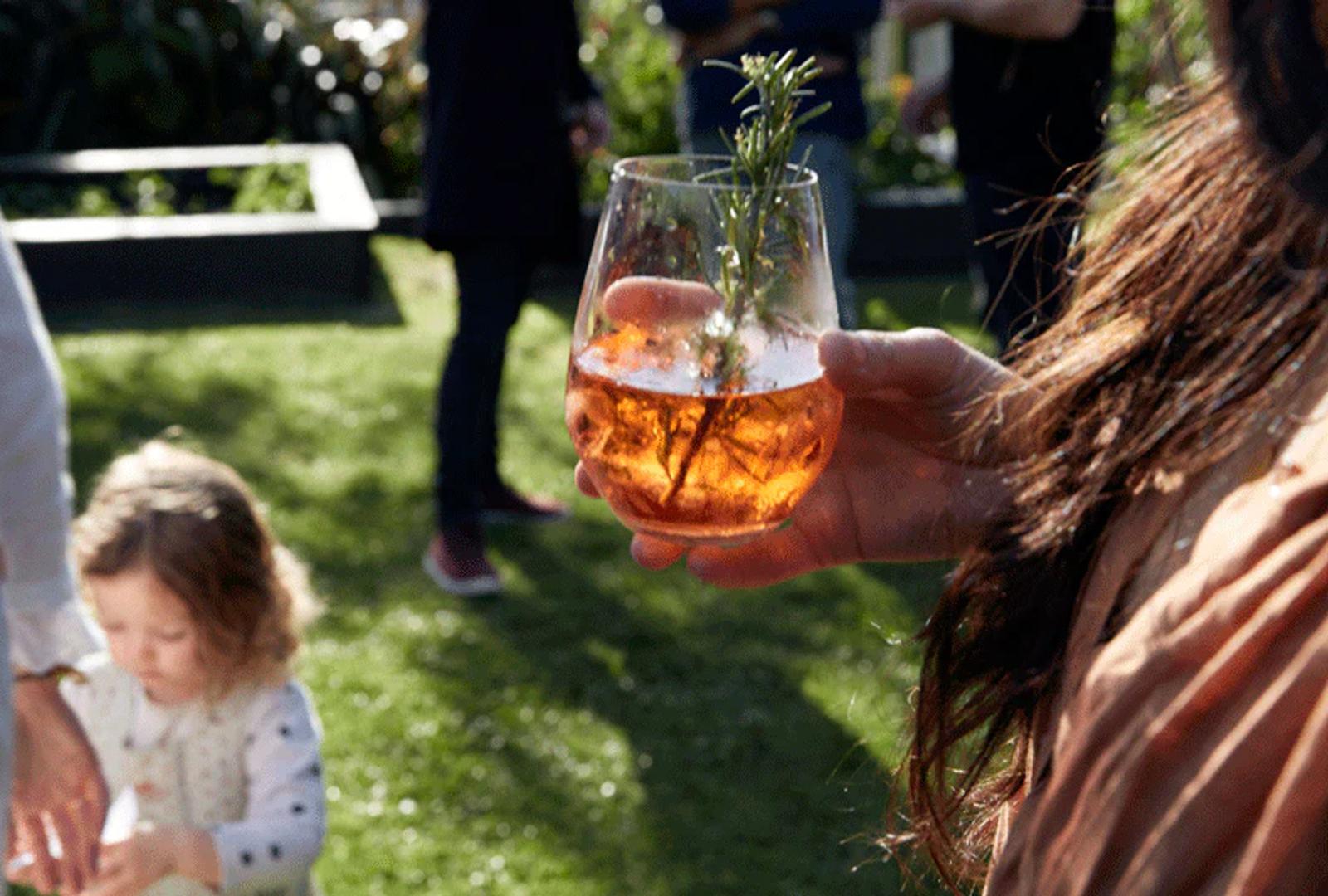 Person with rosemary-garnished red drink at outdoor party. Child playing in background. Sunlit grassy area visible.