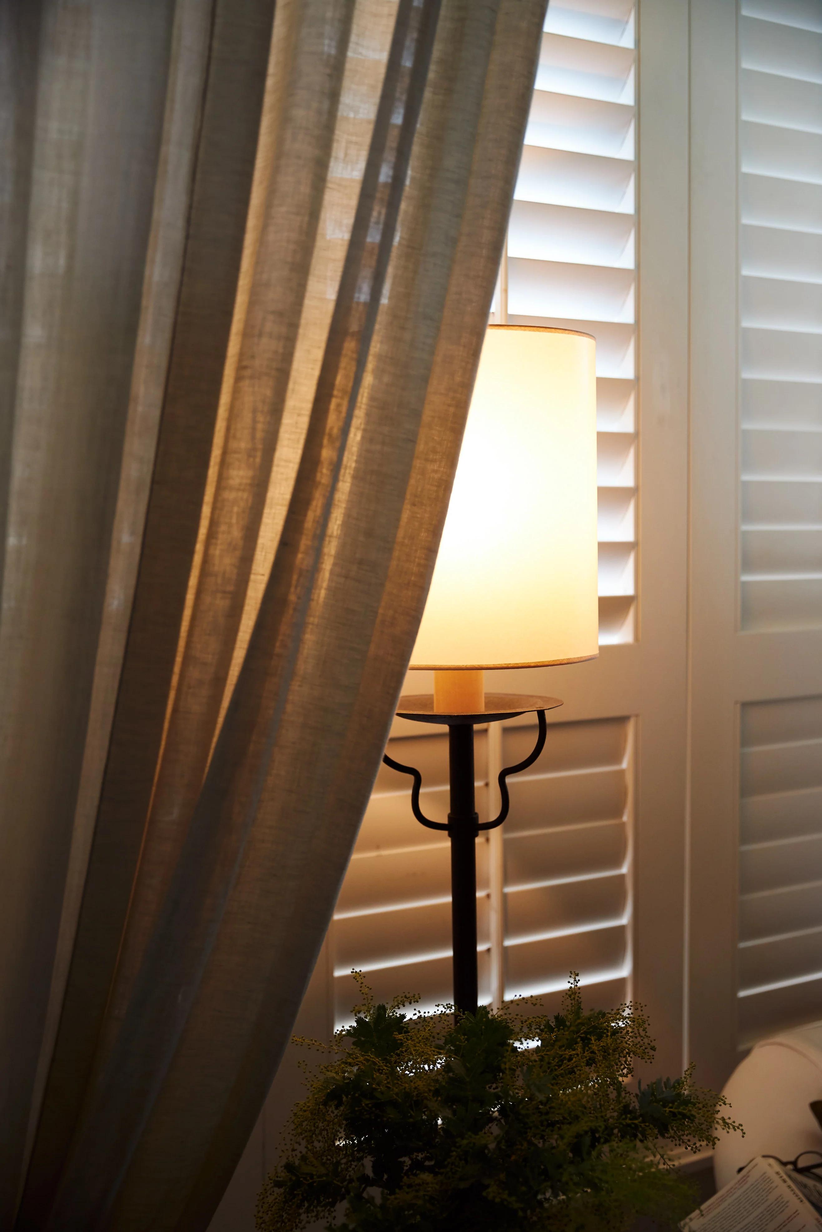 Lit lamp with cylindrical shade on dark stand. Beige curtains, white shutters nearby. Green foliage at base.