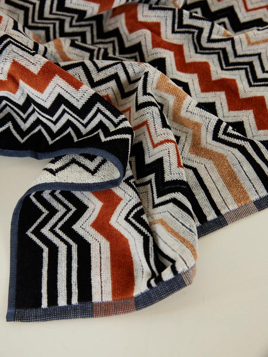 Close-up of a woven blanket with a black, white, rust, and beige zigzag pattern. The slightly wrinkled fabric shows intricate Missoni-style design.