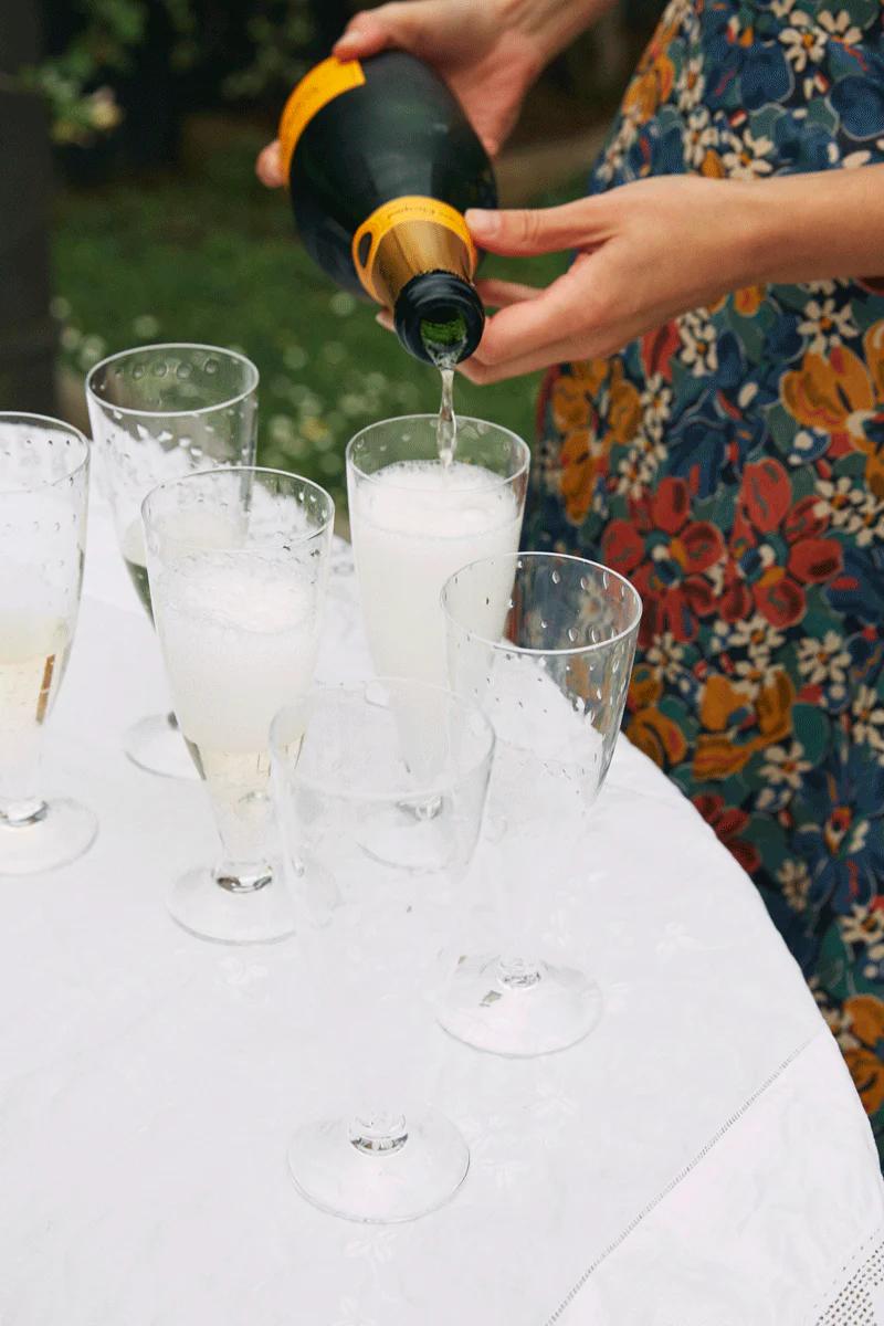 Person in floral dress pours champagne at round table. Multiple glasses create festive atmosphere suitable for holiday gatherings.