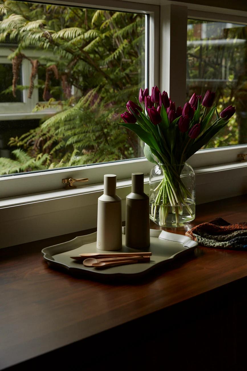 A wooden tray with ceramic bottles and utensils on a kitchen counter. Purple tulips in a vase add colour, with greenery visible through a window.