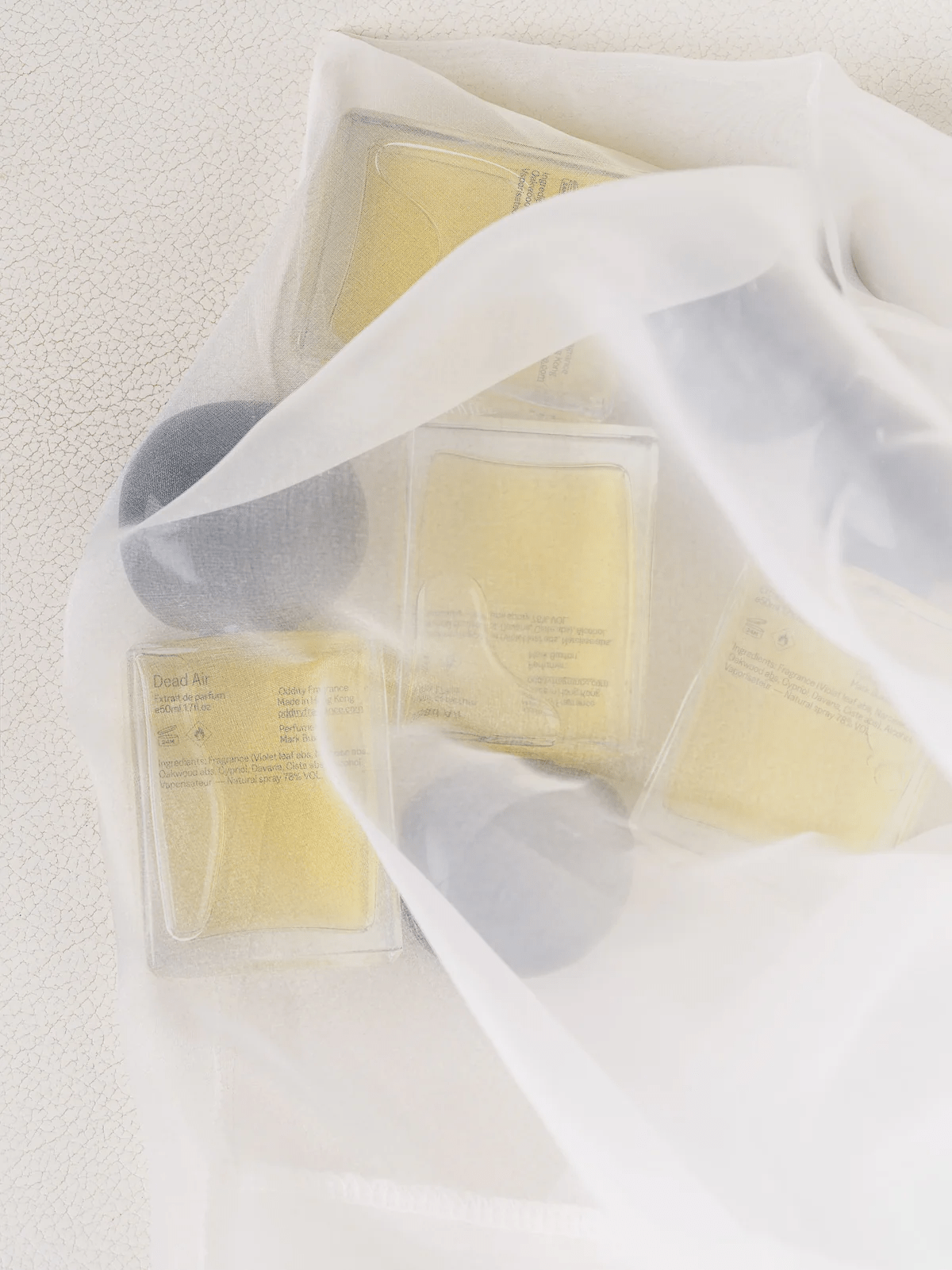 Rectangular perfume bottles with yellow liquid partially covered by sheer white cloth. Two bottles have dark spherical caps.