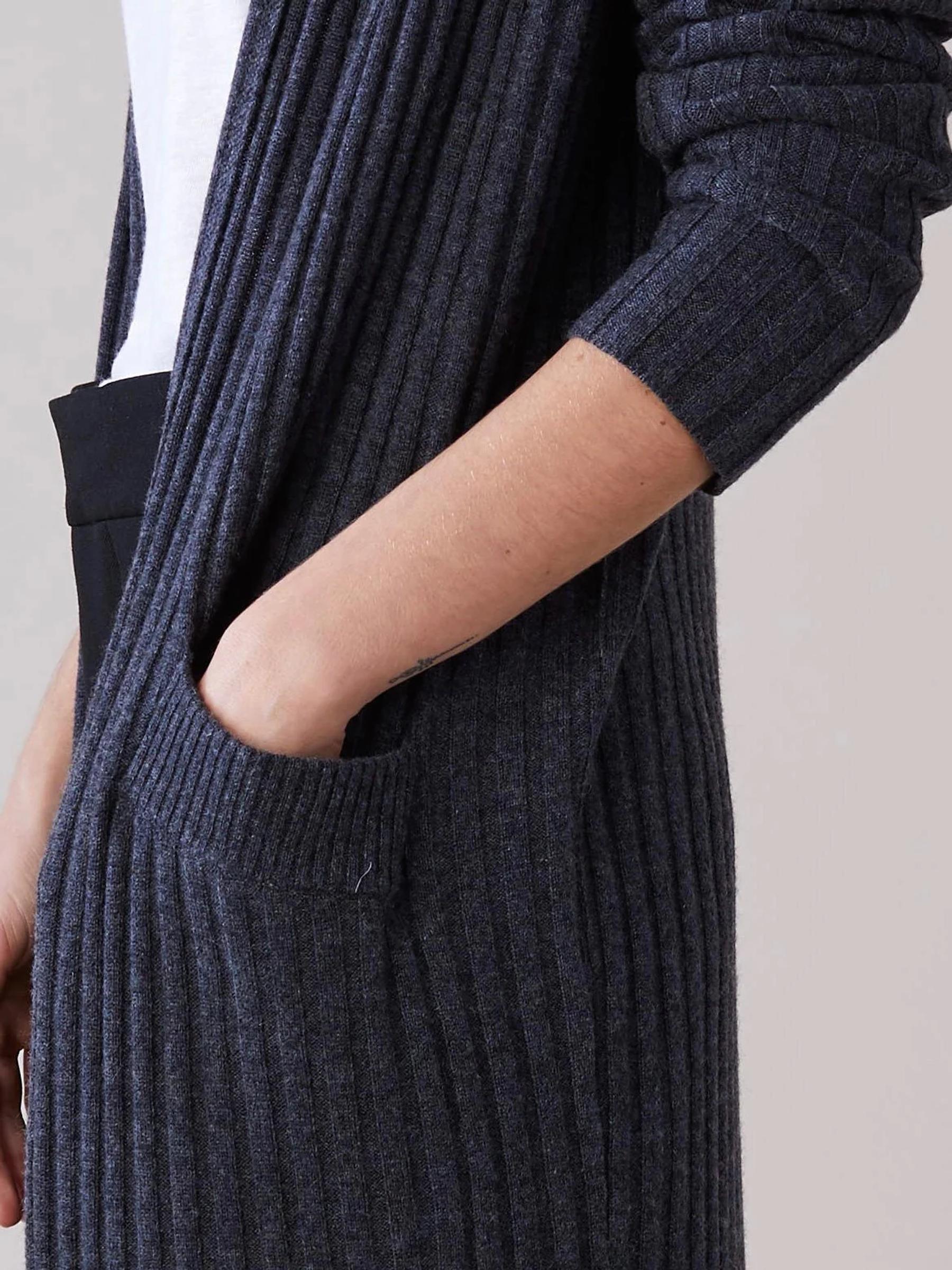 Close-up: Person in long dark gray ribbed cardigan and black pants. Hand in side pocket. Plain light background.