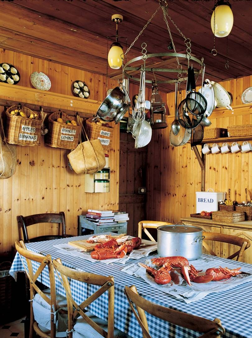 Jura kitchen with hanging pots and wicker baskets. Table set with lobsters, pot, and books. Blue checkered tablecloth.