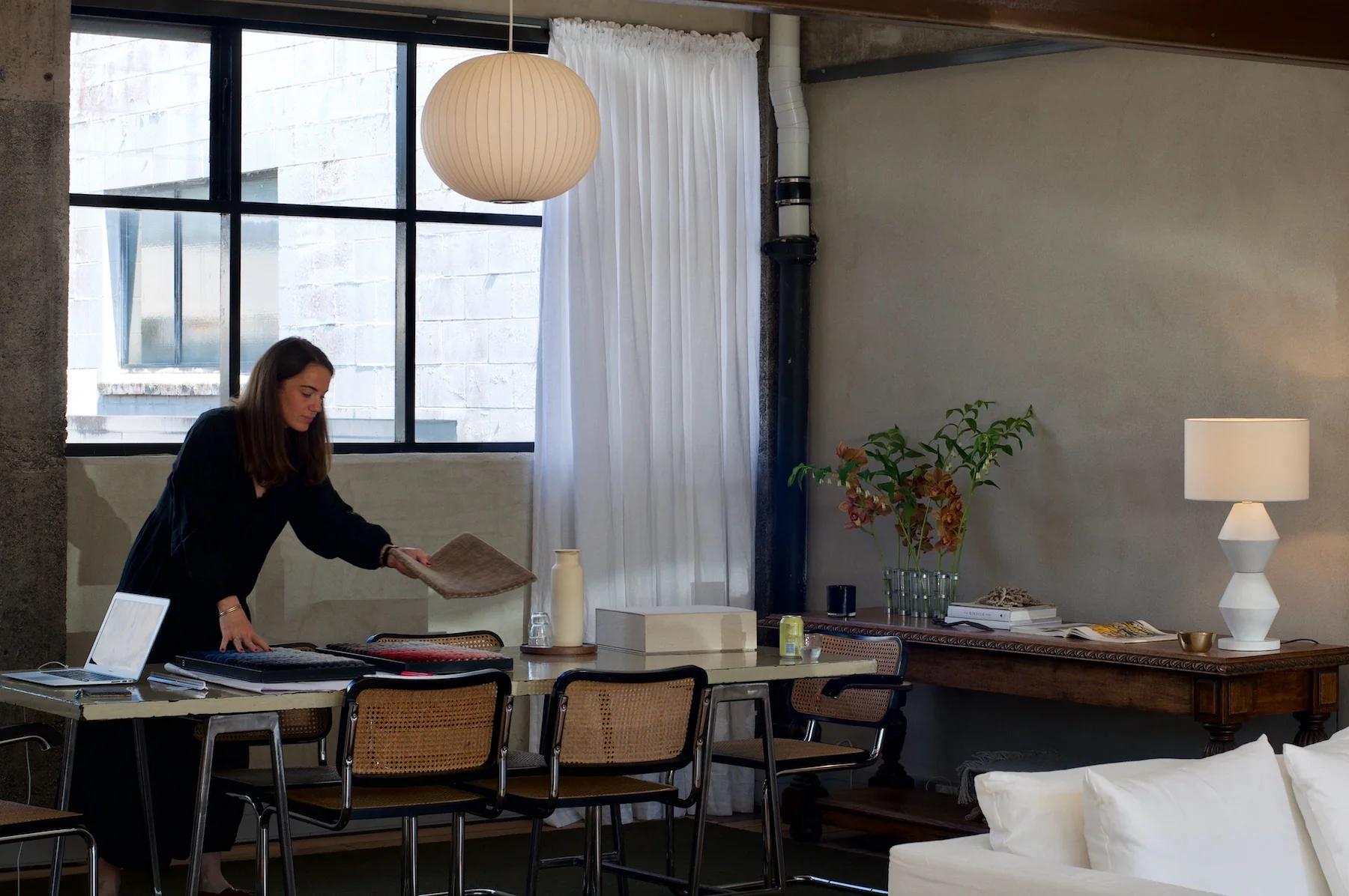 Woman at table with interior samples in modern room. Contemporary furniture, window, and plant visible.