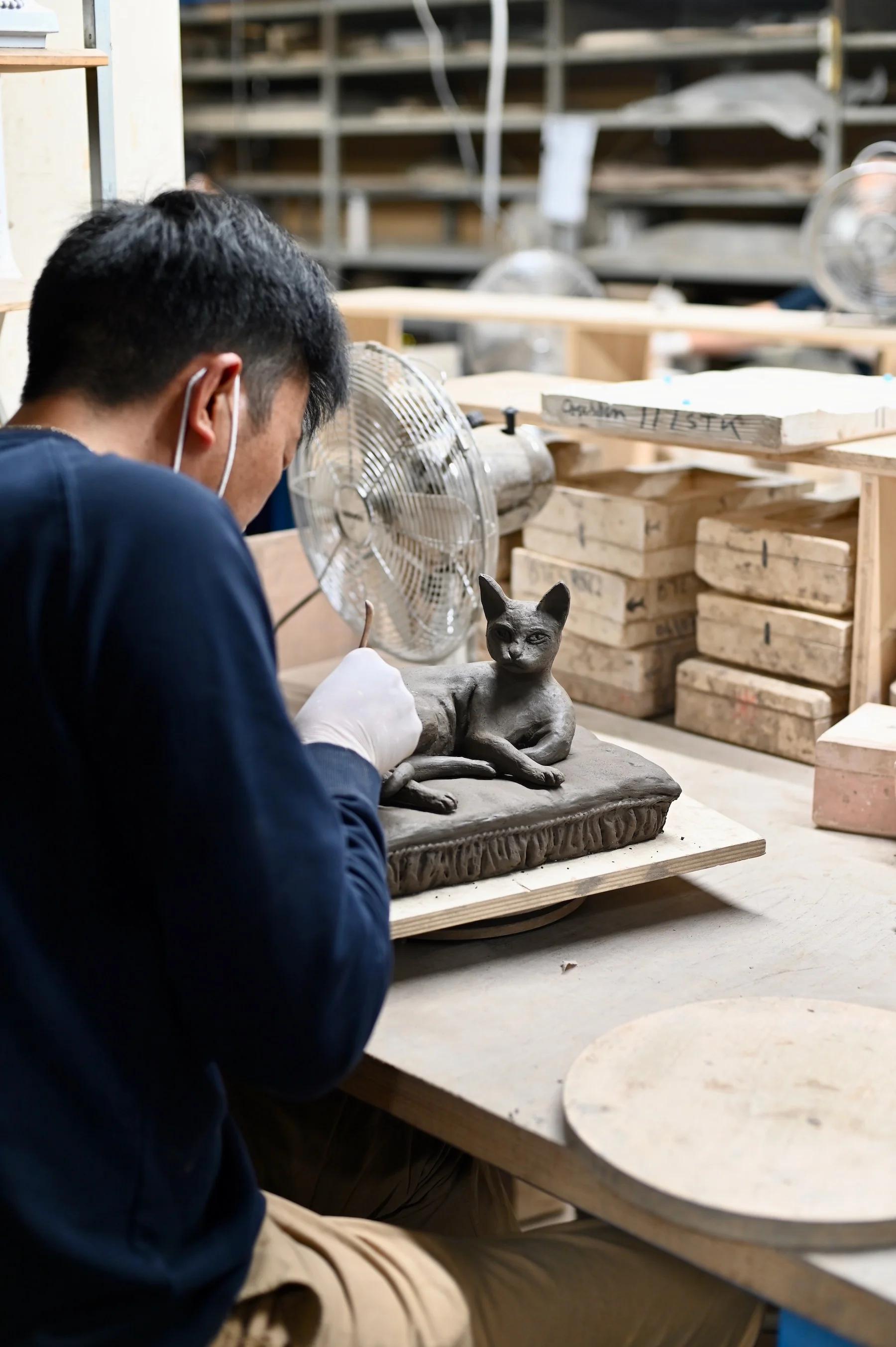 Person wearing mask and gloves sculpting cat figurine on cushion. Cluttered workspace with wooden boxes, shelves, and fan in background.