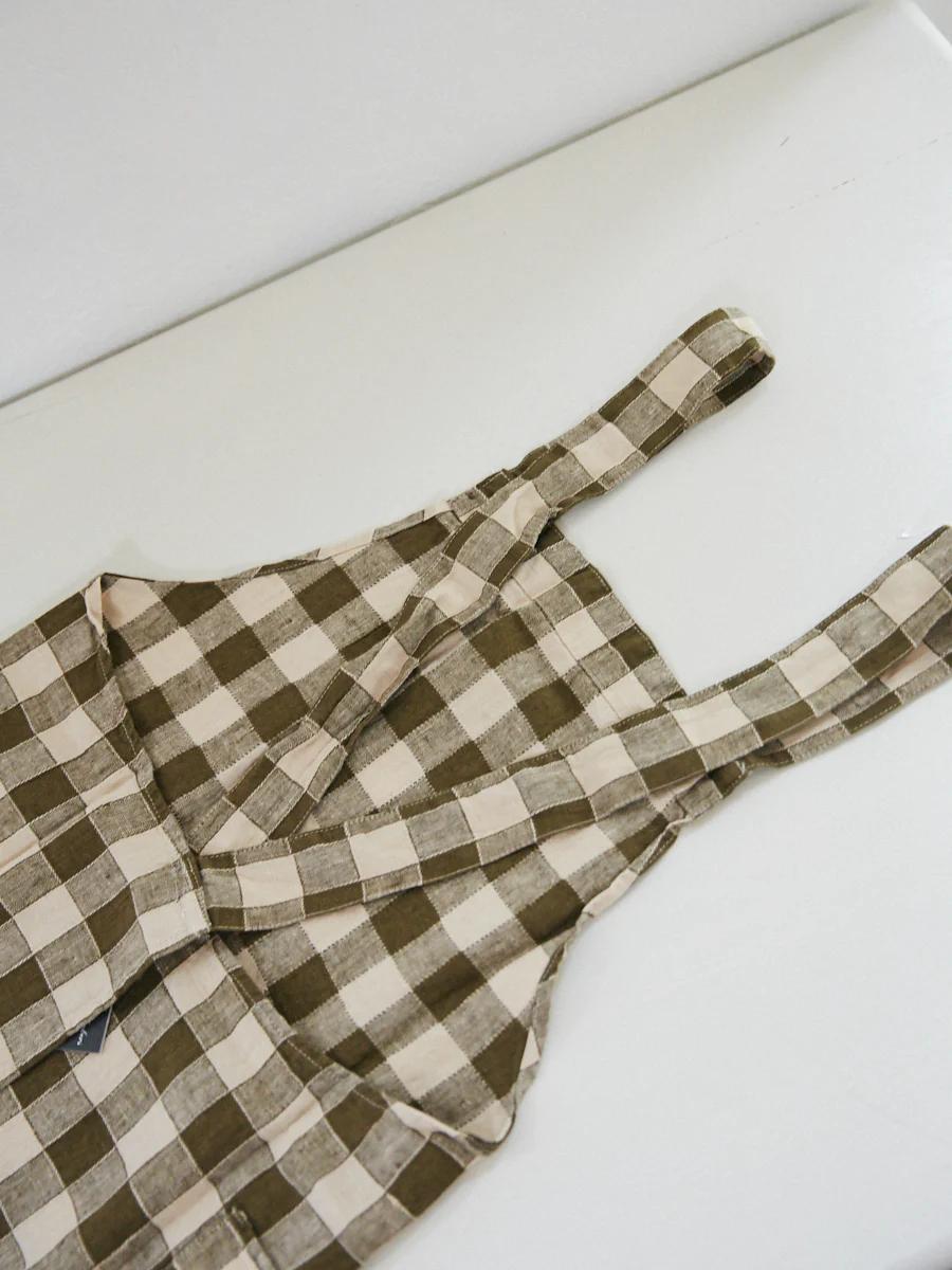 Checkered cloth apron with brown and white squares. Two straps, front pocket. Laid flat on white surface.