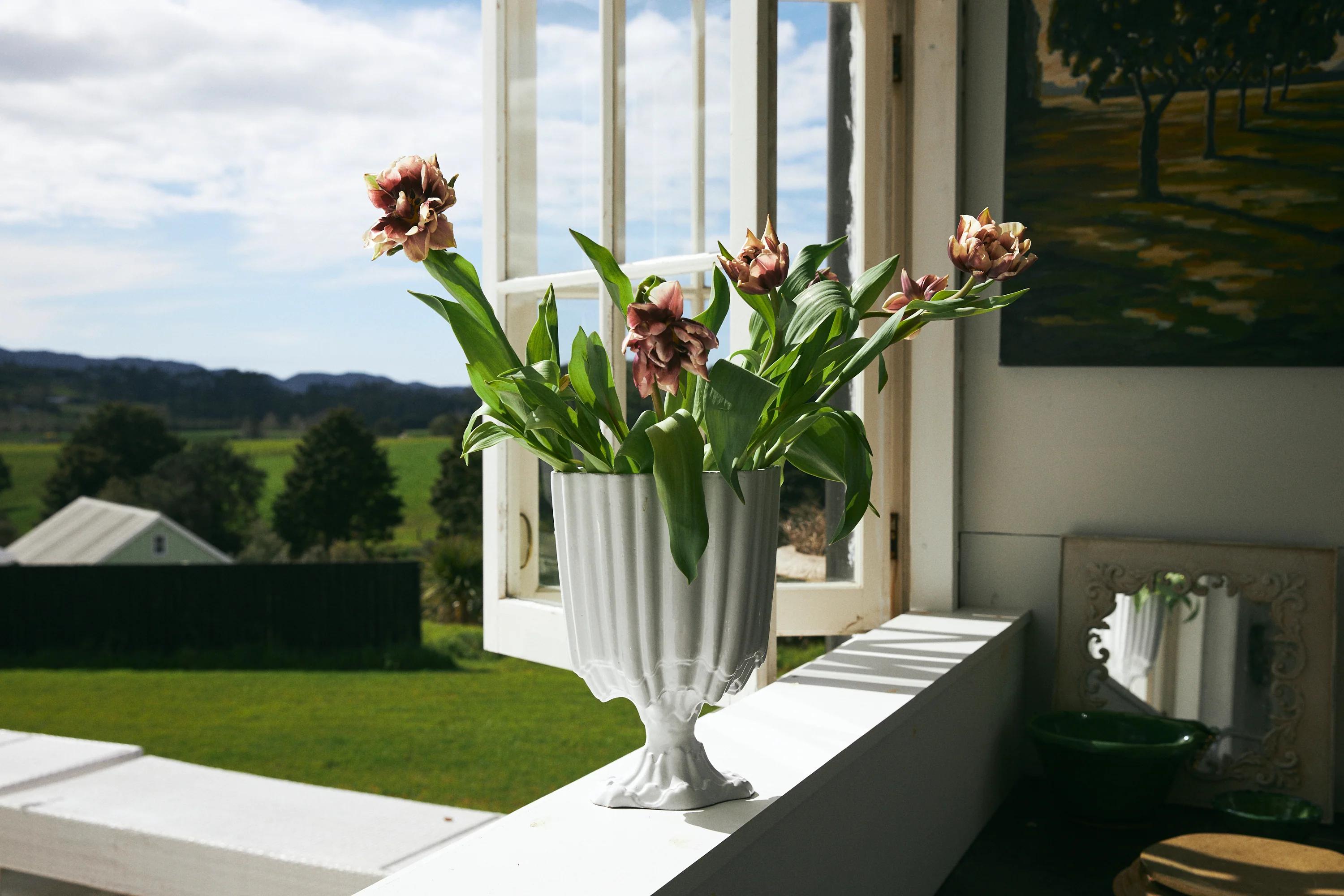 Pink potted plant on sunny windowsill. Open window shows countryside view. Painting and Astier de Villatte tableware on adjacent wall.