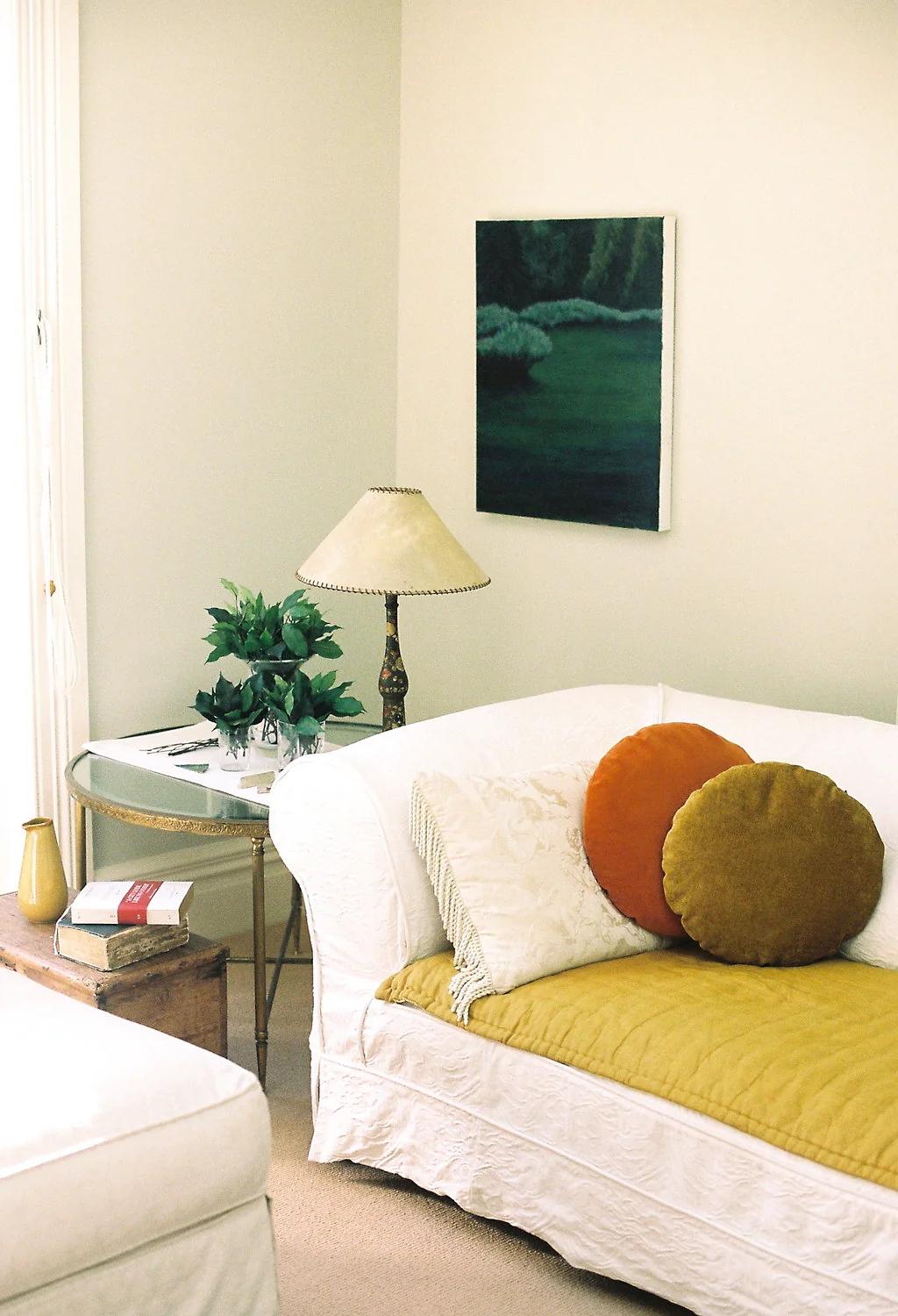 Cosy living room with white sofa, golden and orange KLAY cushions and quilt. Side table with plant, lamp, and books nearby. Framed art and wooden chest complete scene.