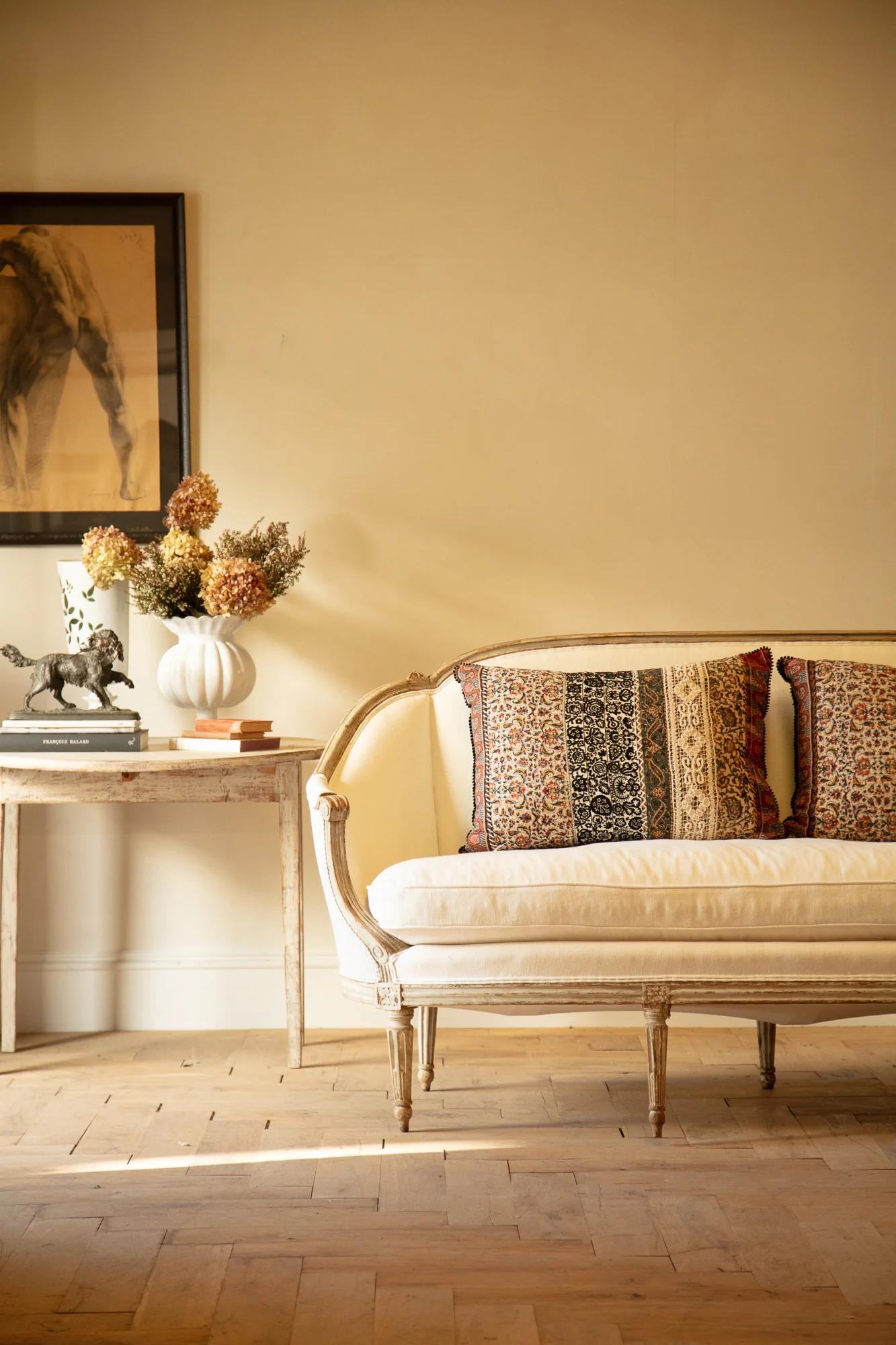 Living room with cushioned wooden bench. Side table with books and grapes. Floor cushions, patterned rug, and floral artwork create British ambience.