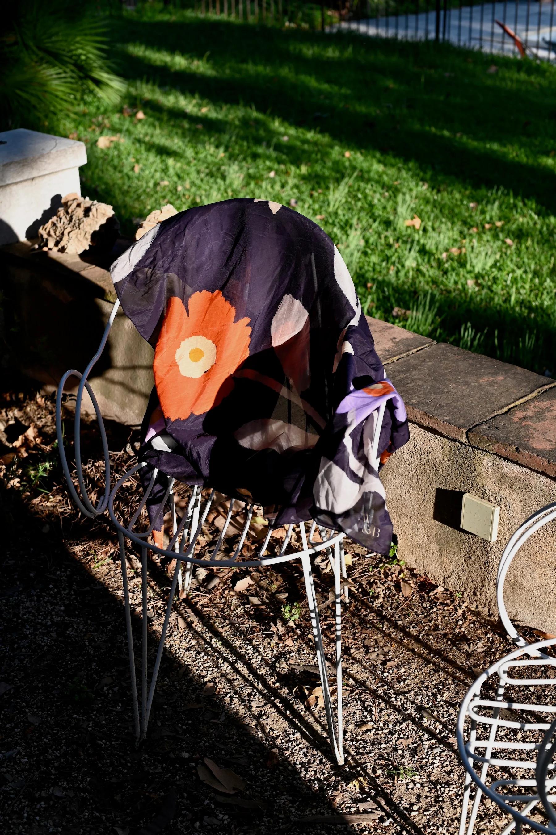 Wire-frame chair with curved armrests on gravel path. Black cloth with orange flower draped over chair. Grassy lawn and stone wall in background.