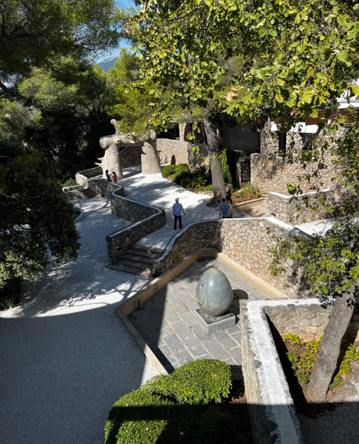 Curved garden path with stone walls and sculptures. People walking, observing. Trees and bushes alongside.