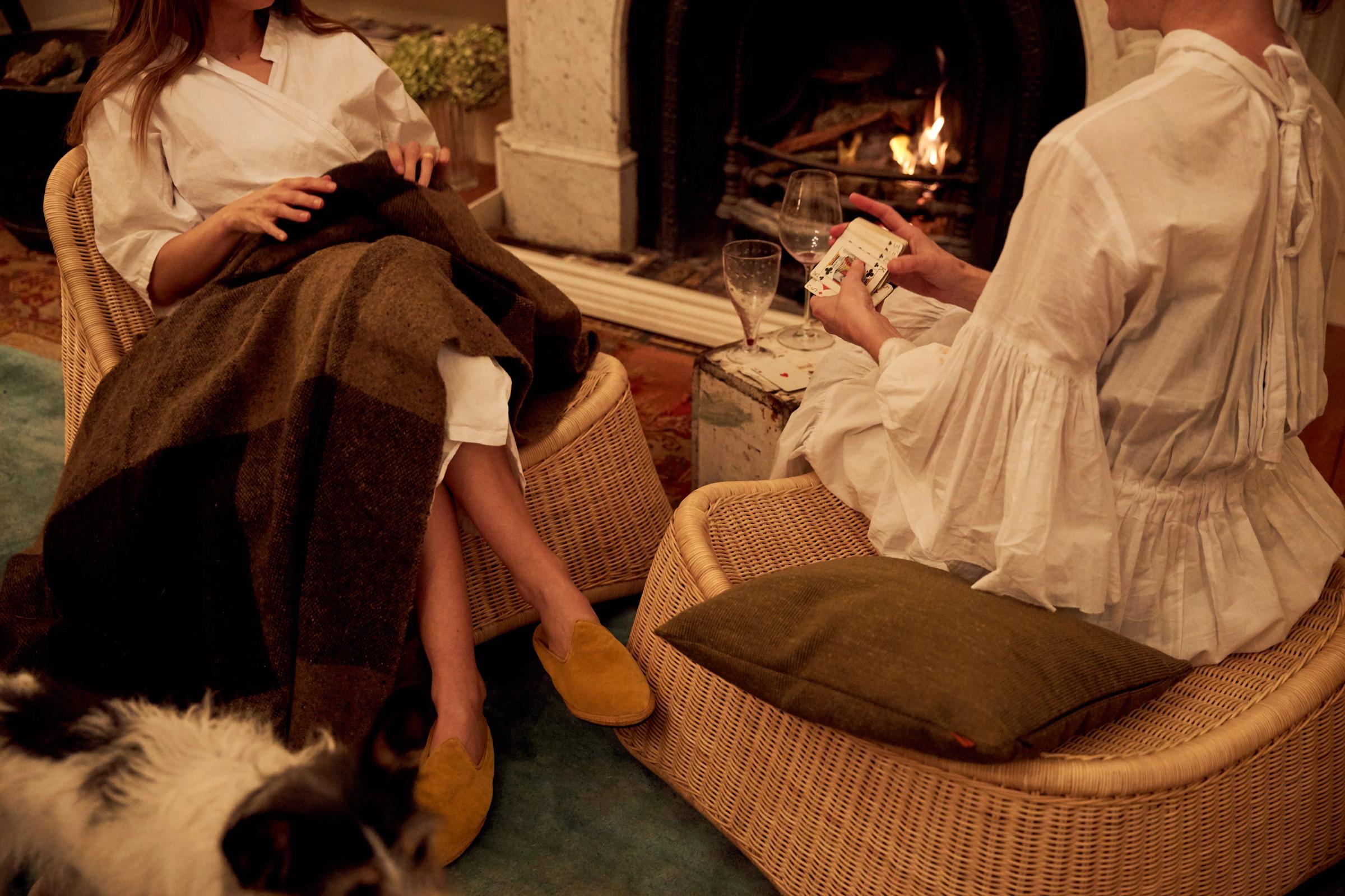 Two people dressed in white sit on wicker chairs beside a fire. One has a brown blanket, the other sits beside a brown cushion whilst shuffling a deck of cards.
