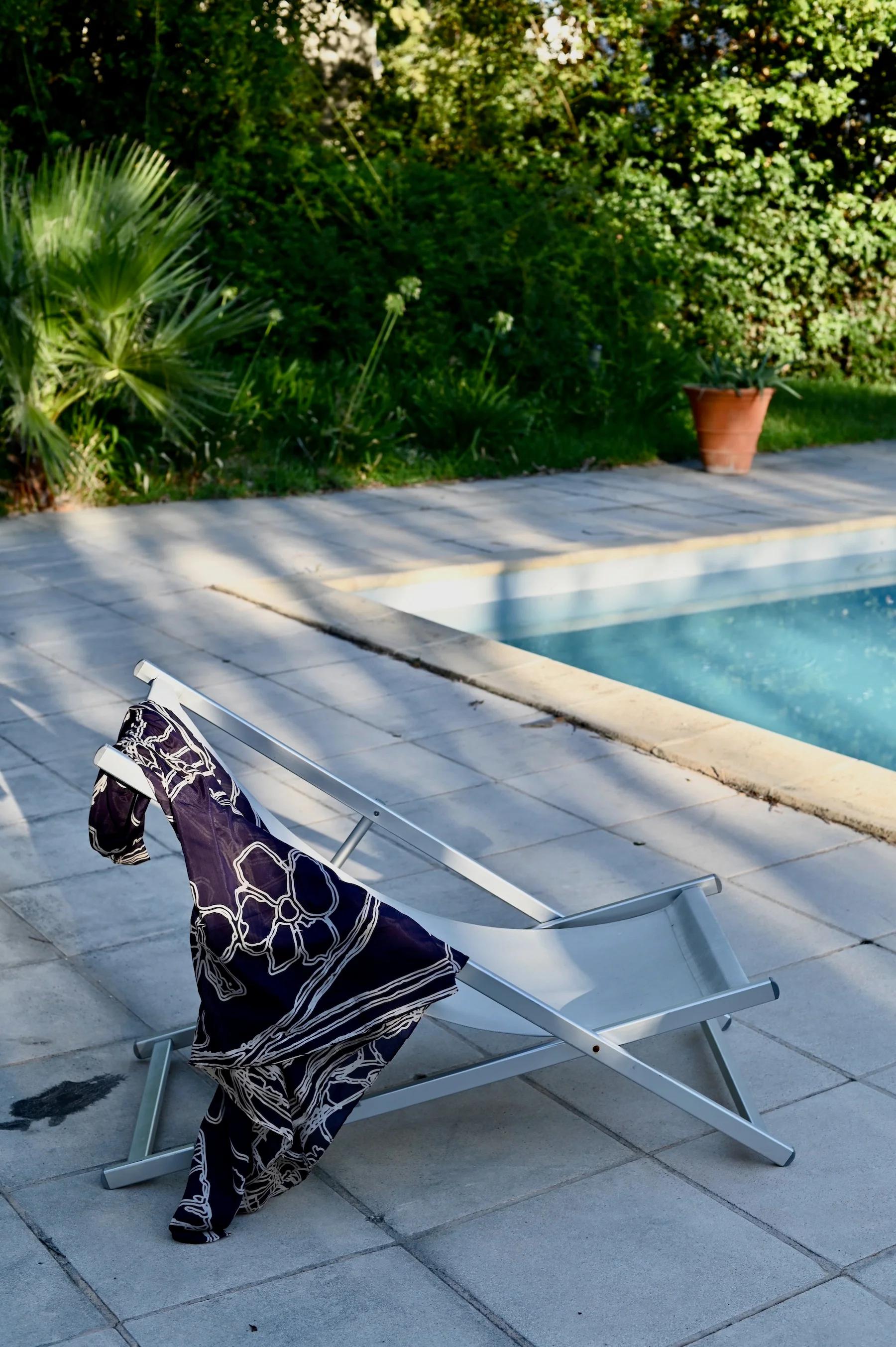 Sun lounger with patterned fabric by swimming pool. Clear blue water contrasts with tiled poolside. Lush greenery and potted plant in background.