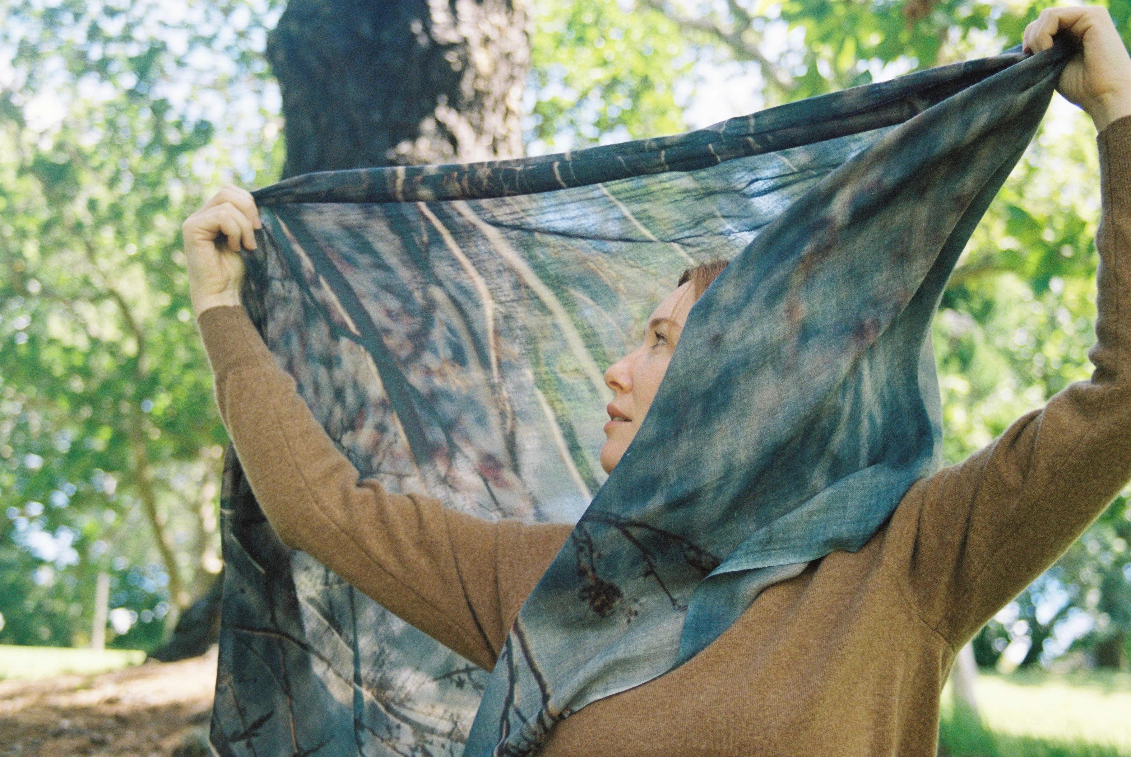 Person in brown sweater holding translucent, branch-patterned scarf overhead. Sunlit forest background with large tree trunk.