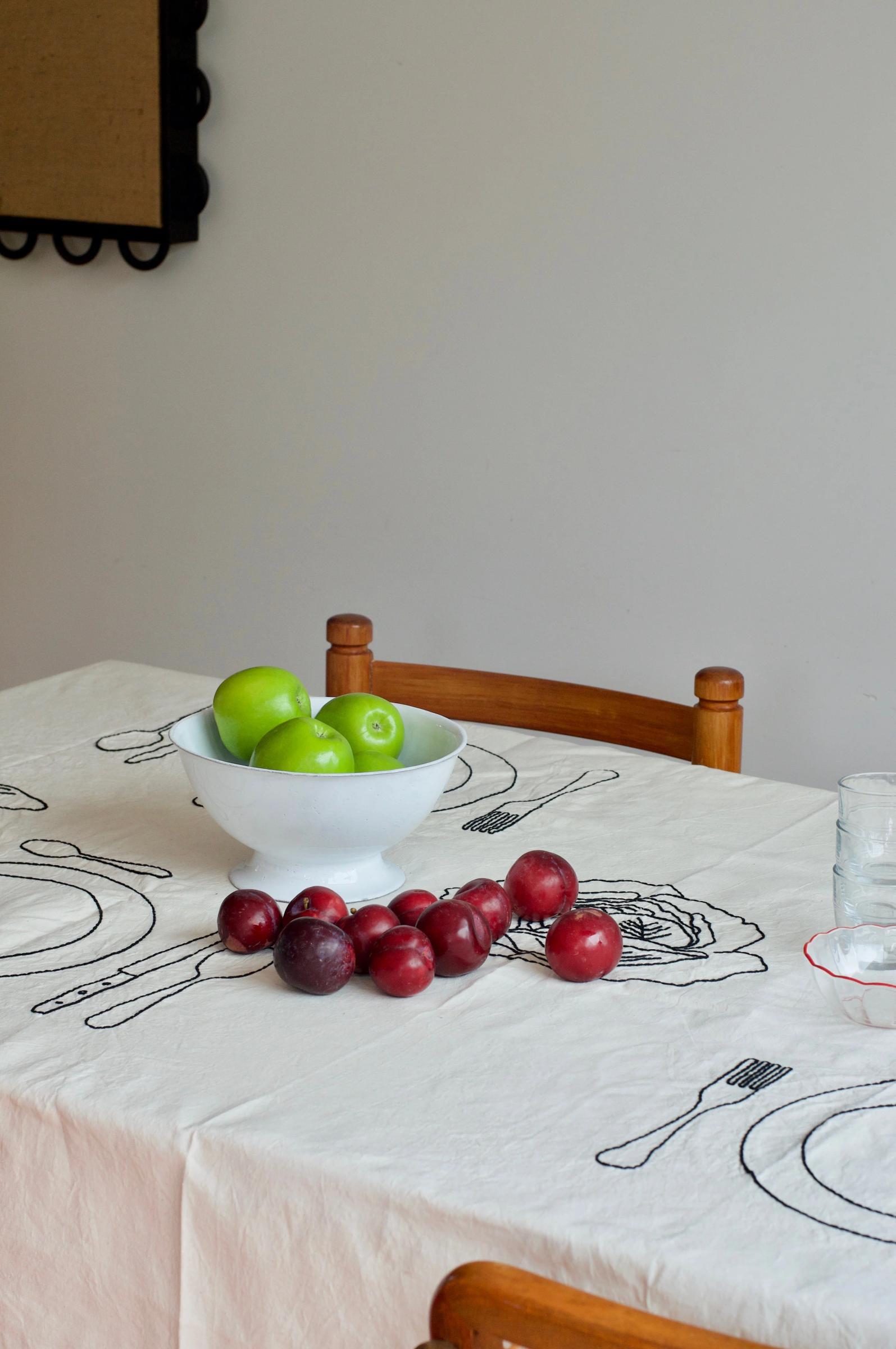 Table with embroidered linens, white bowl of green apples, red plums scattered in front. Two wooden chairs, glass tumbler nearby.