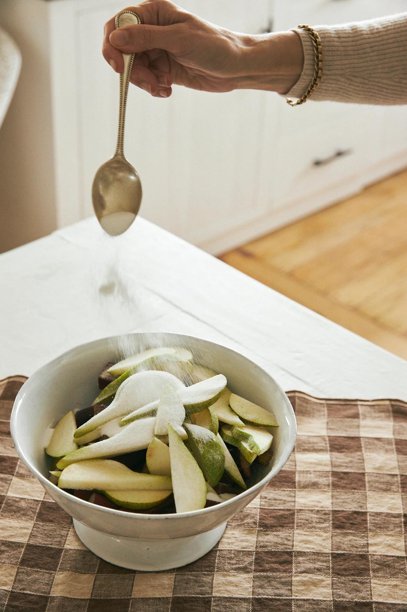 Hand sprinkling sugar on sliced pears in white bowl. Checkered cloth, white table. Cabinet and wooden floor visible.