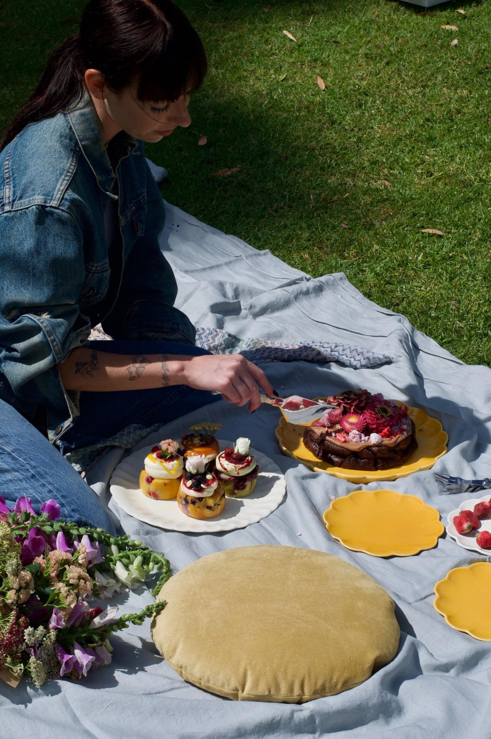 A person arranges cakes and pastries from The Caker bakery on a picnic blanket, surrounded by flowers and a yellow cushion, set against green grass and fallen petals.\