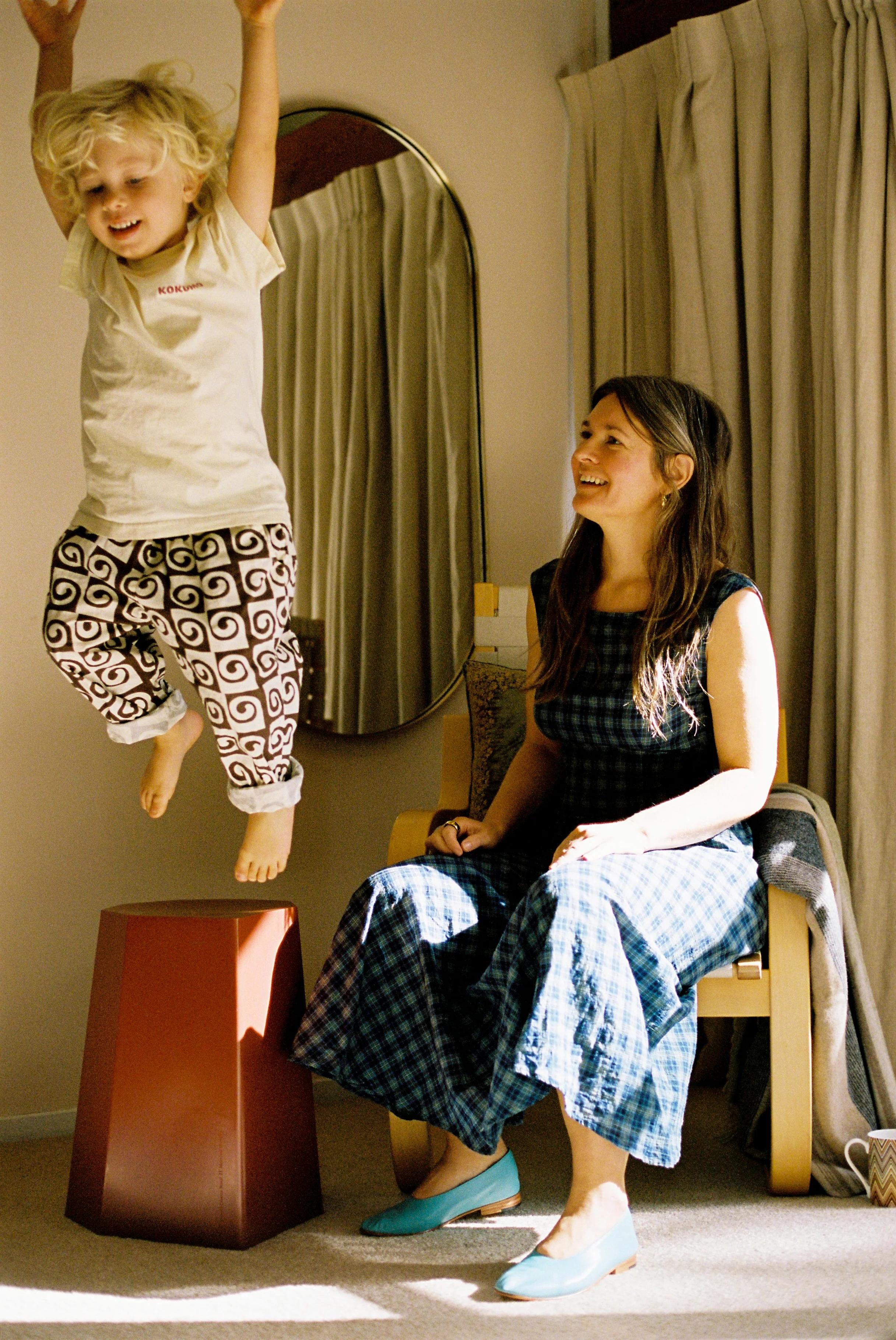 Blonde child jumping off red stool. Woman in blue dress watching from chair. Midcentury home setting.