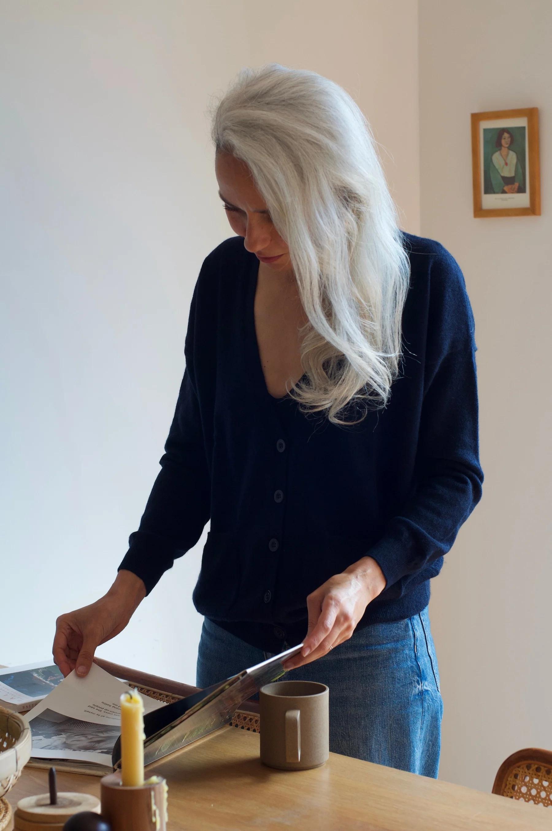 White-haired person at table with magazine. Wearing navy cardigan and jeans. Background shows mug, candle, and framed picture.