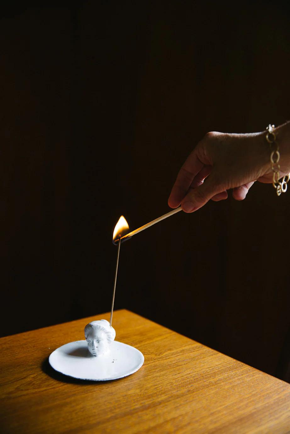 Hand with bracelet lighting head-shaped candle on white saucer. Dark background emphasizing flame.