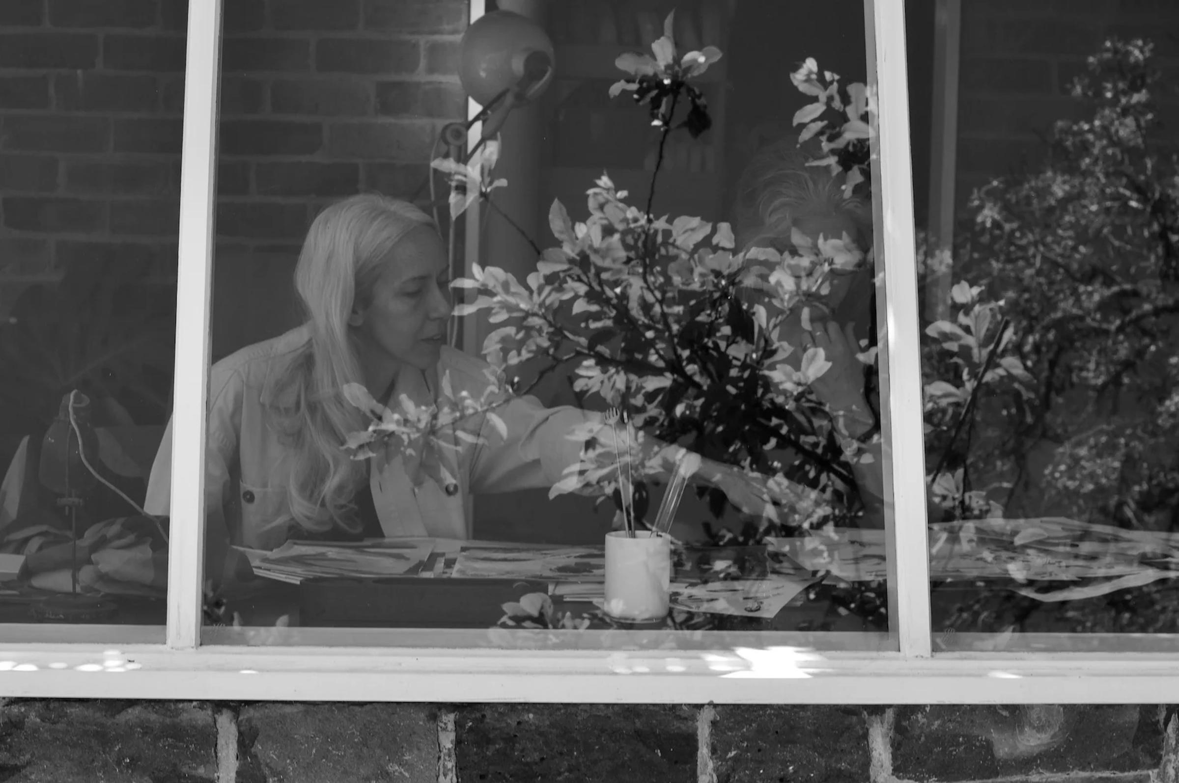 Black and white: Woman at table seen through window. Reaching across table, flower reflections in window.