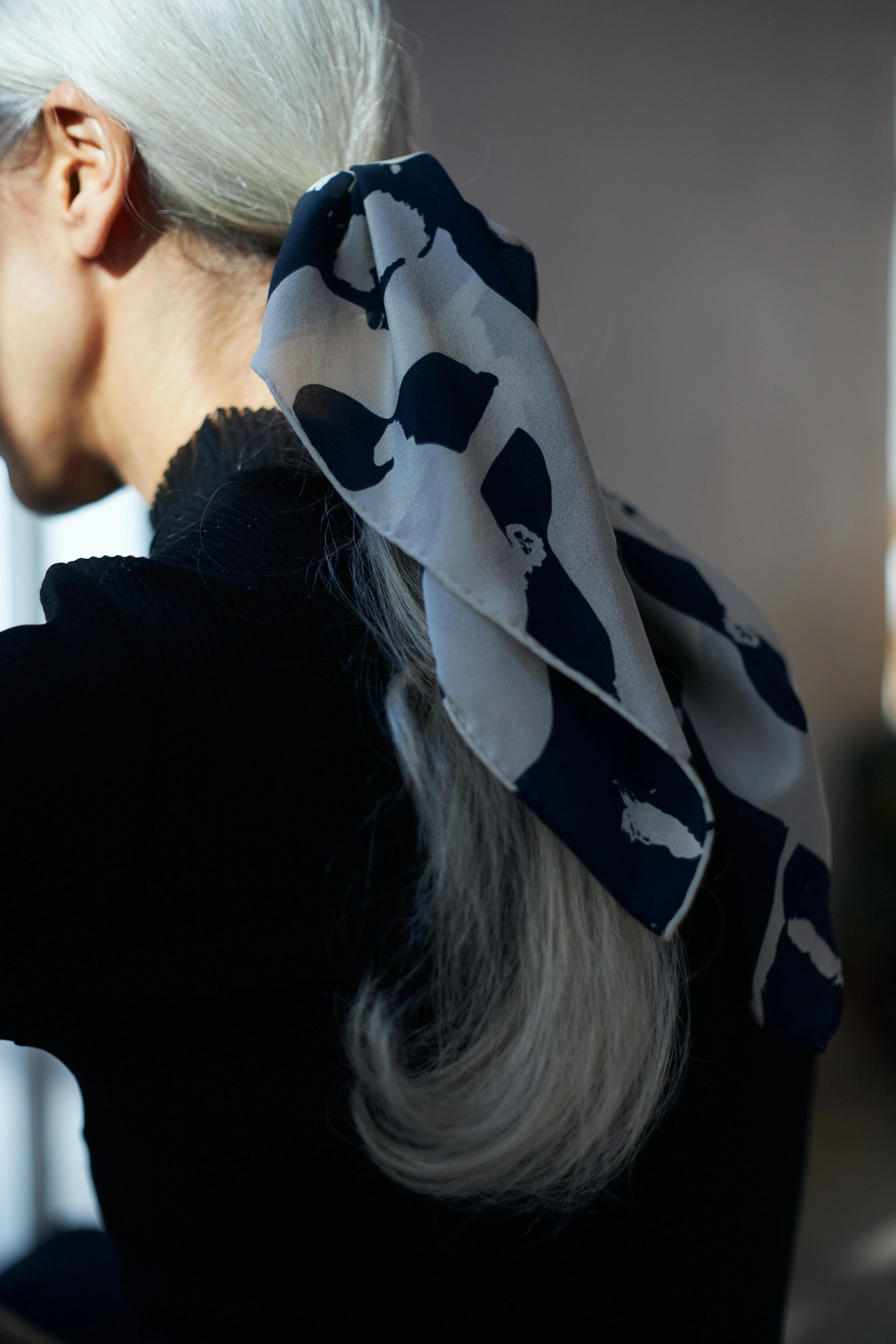 A person with long, silver hair is seen from behind, wearing a high neck black top. Their hair is tied back with a large navy blue and white patterned scarf from Walker & Bing's latest collection, securing a low ponytail. The background is out of focus, emphasising the contrast between the dark clothing and the textile design accessory.