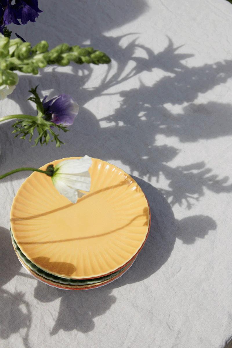 Three Rachel Carley plates are stacked on a light tablecloth. A delicate white flower rests on the yellow plate, with purple flowers and greenery visible in the background.