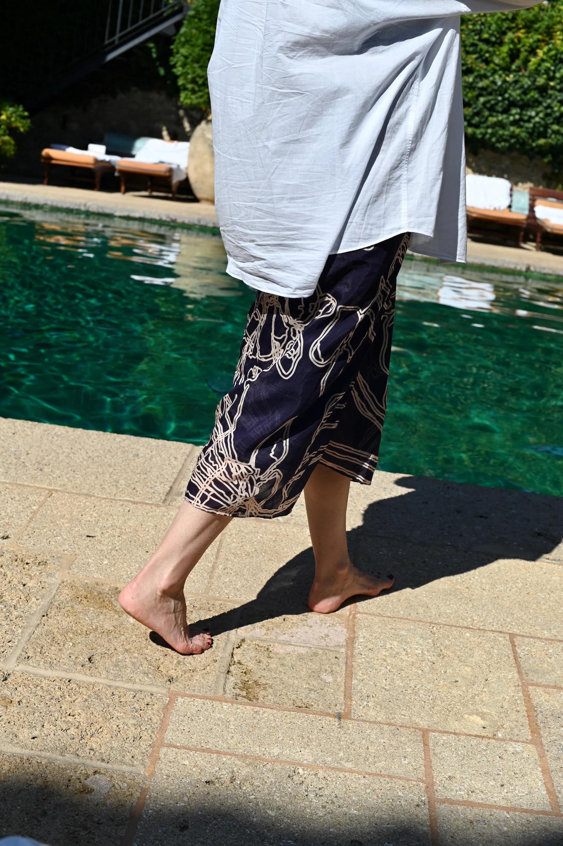 Person walking barefoot by pool in navy pants with white pattern and oversized light blue shirt. Clear water and lounge chairs in background.