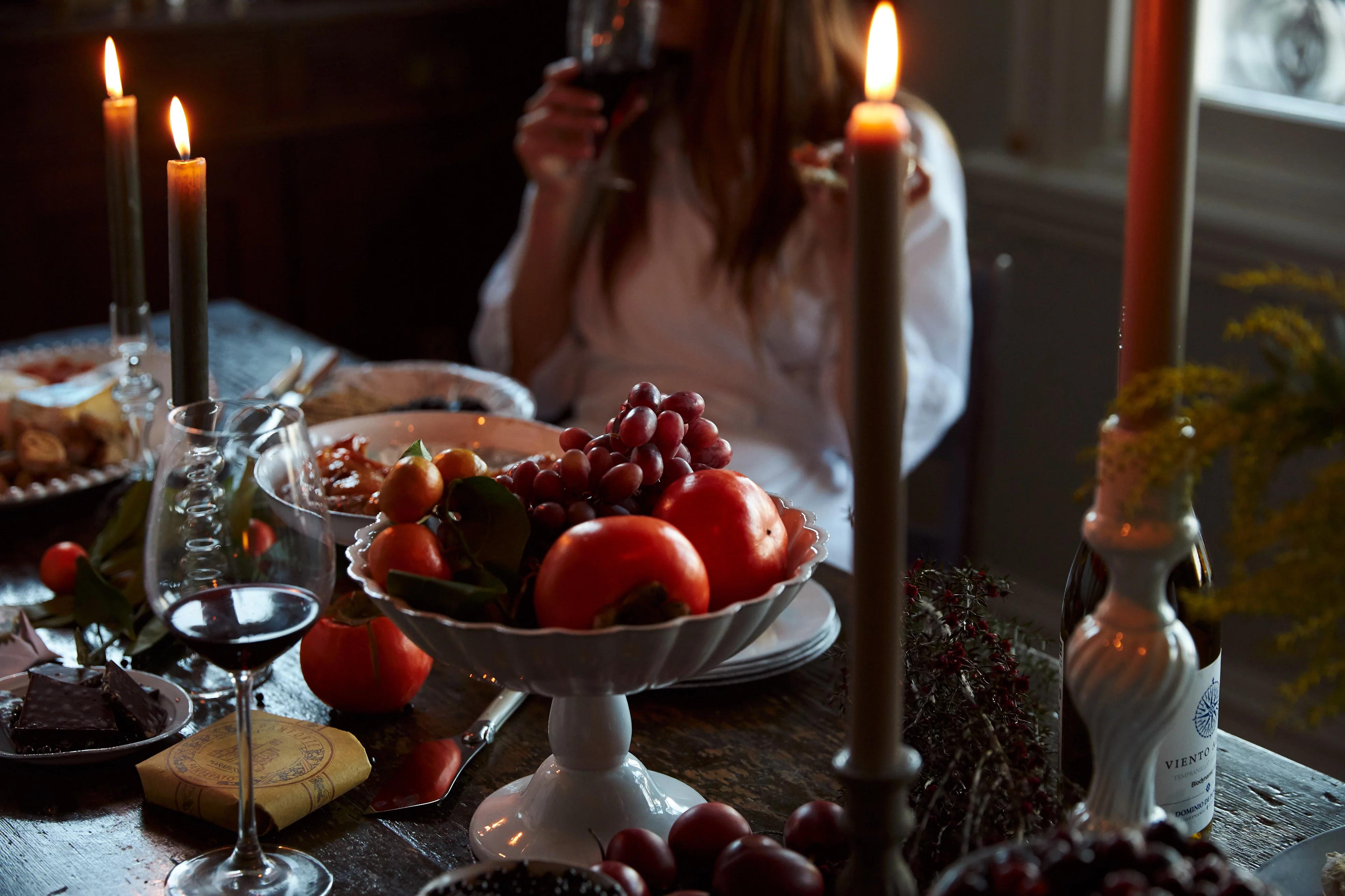 Candlelit table with wine, fruit bowl, and food. Person drinking wine in blurred background.