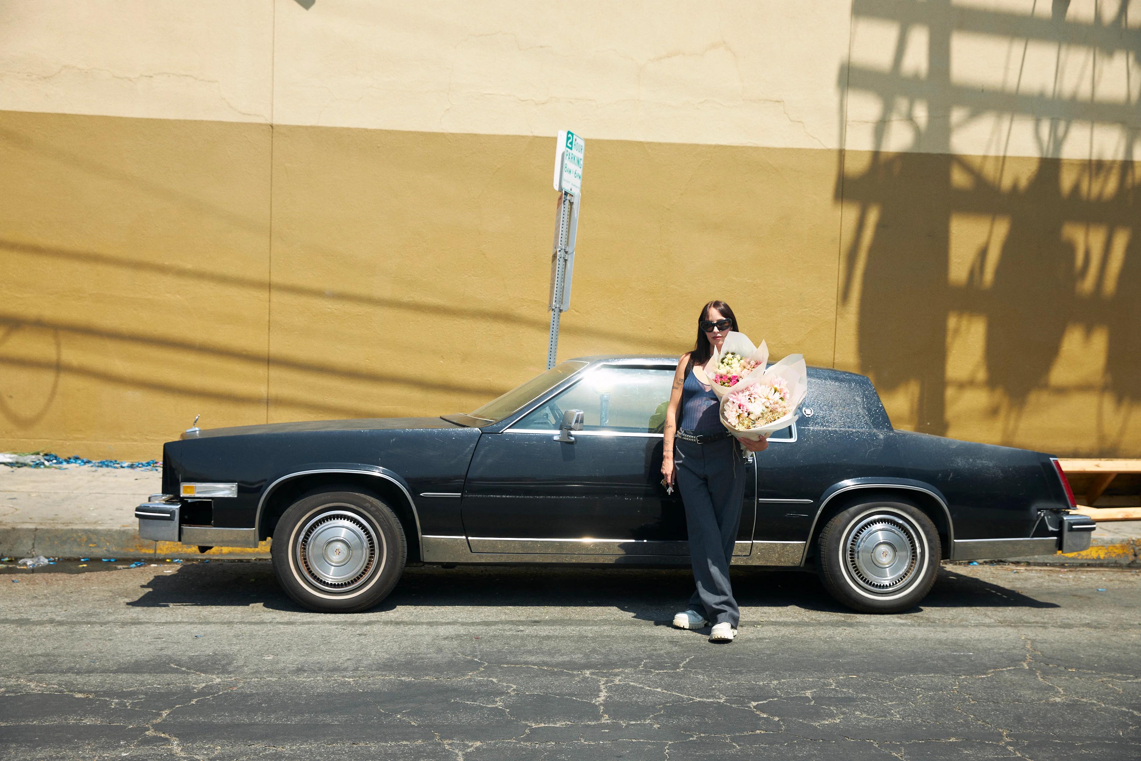 Person in sunglasses by vintage black car. Holding large flower bouquet, dressed in dark outfit. Plain wall and street sign behind.