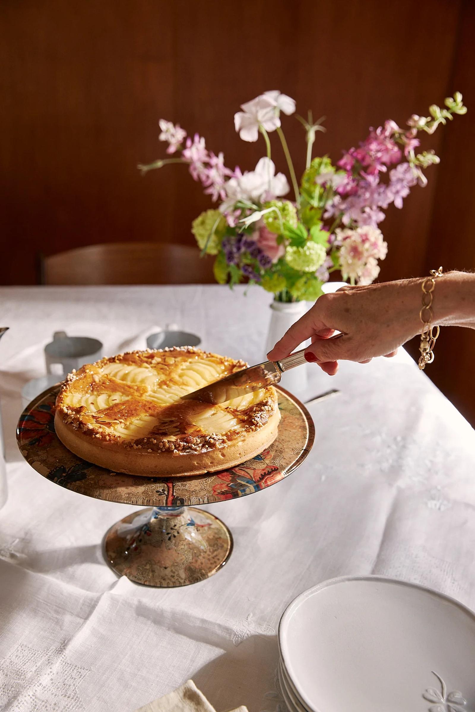 Woman cuts into a pear tart placed on a glass cake stand with flowers in the background