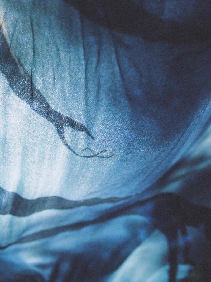 Close-up of flowing fabric in shades of blue and indigo with abstract, branch-like lines. Soft light highlights the wrinkles, creating a shadowy, textured effect.