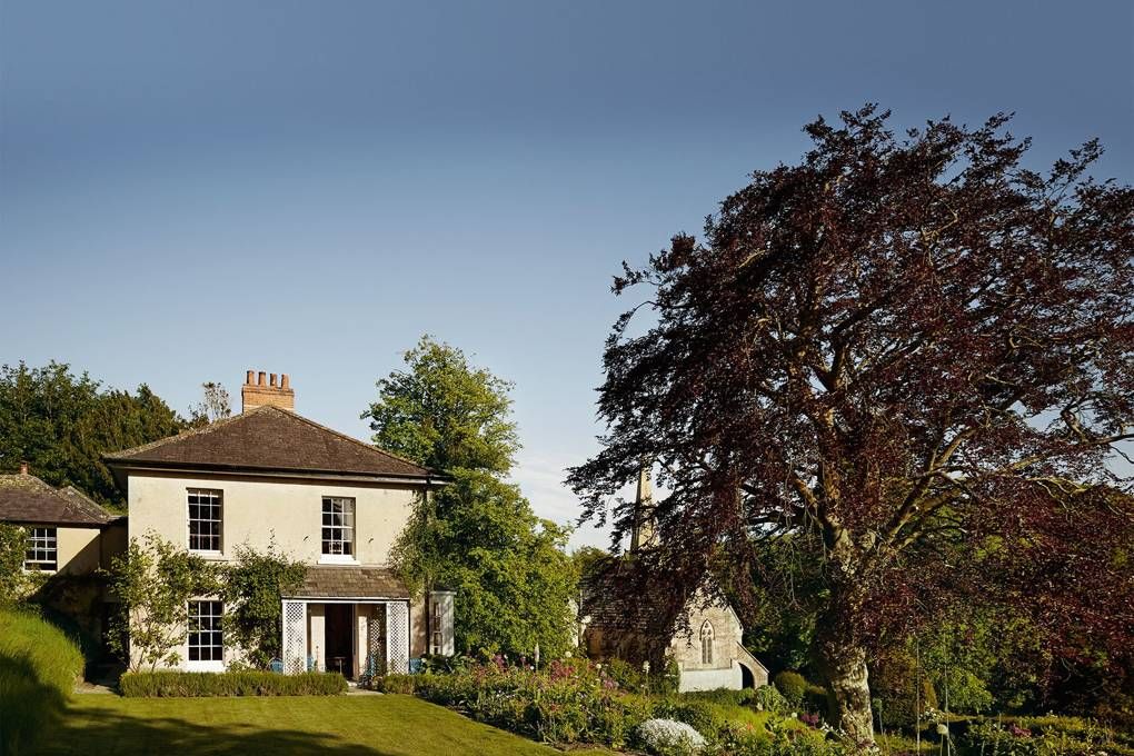 Cream house with steep roof in lush garden. Large tree with reddish-brown leaves in foreground, blue sky behind.