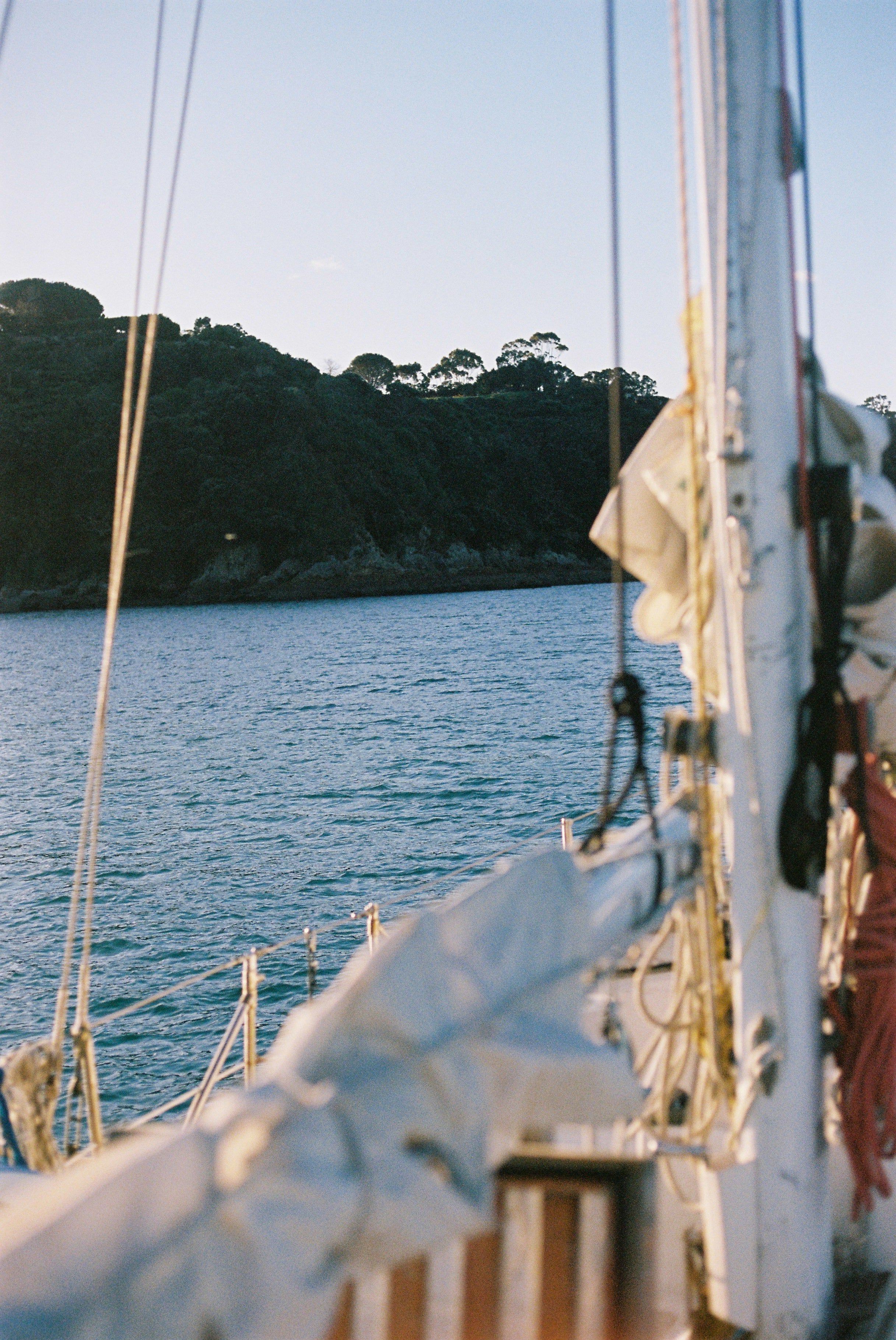 Sailboat deck view showing mast, rigging, and sails. Calm waters and tree-covered hill in background.