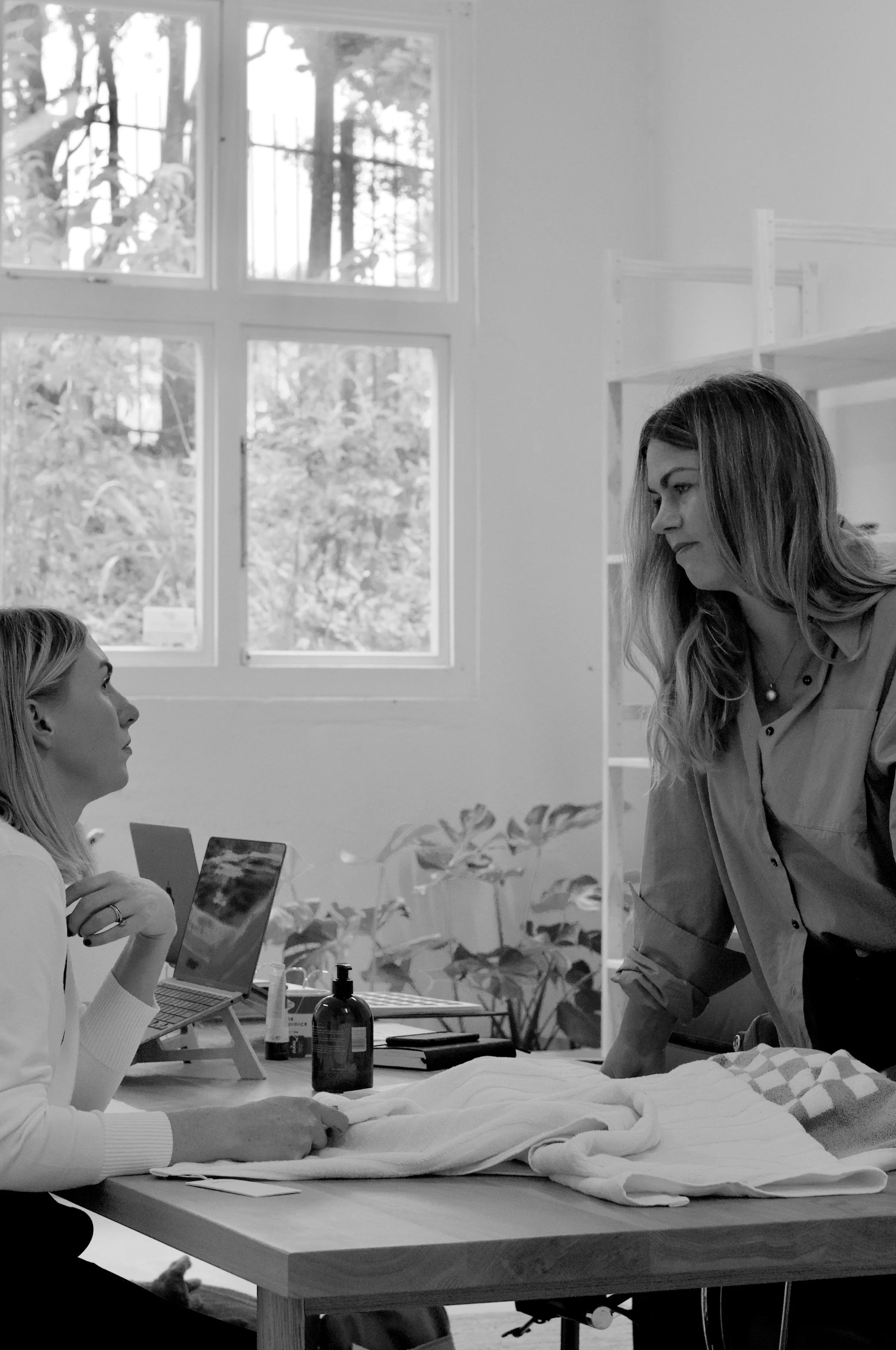 Black and white photo of two women working in office. One seated at desk, one standing. Large windows, natural light.