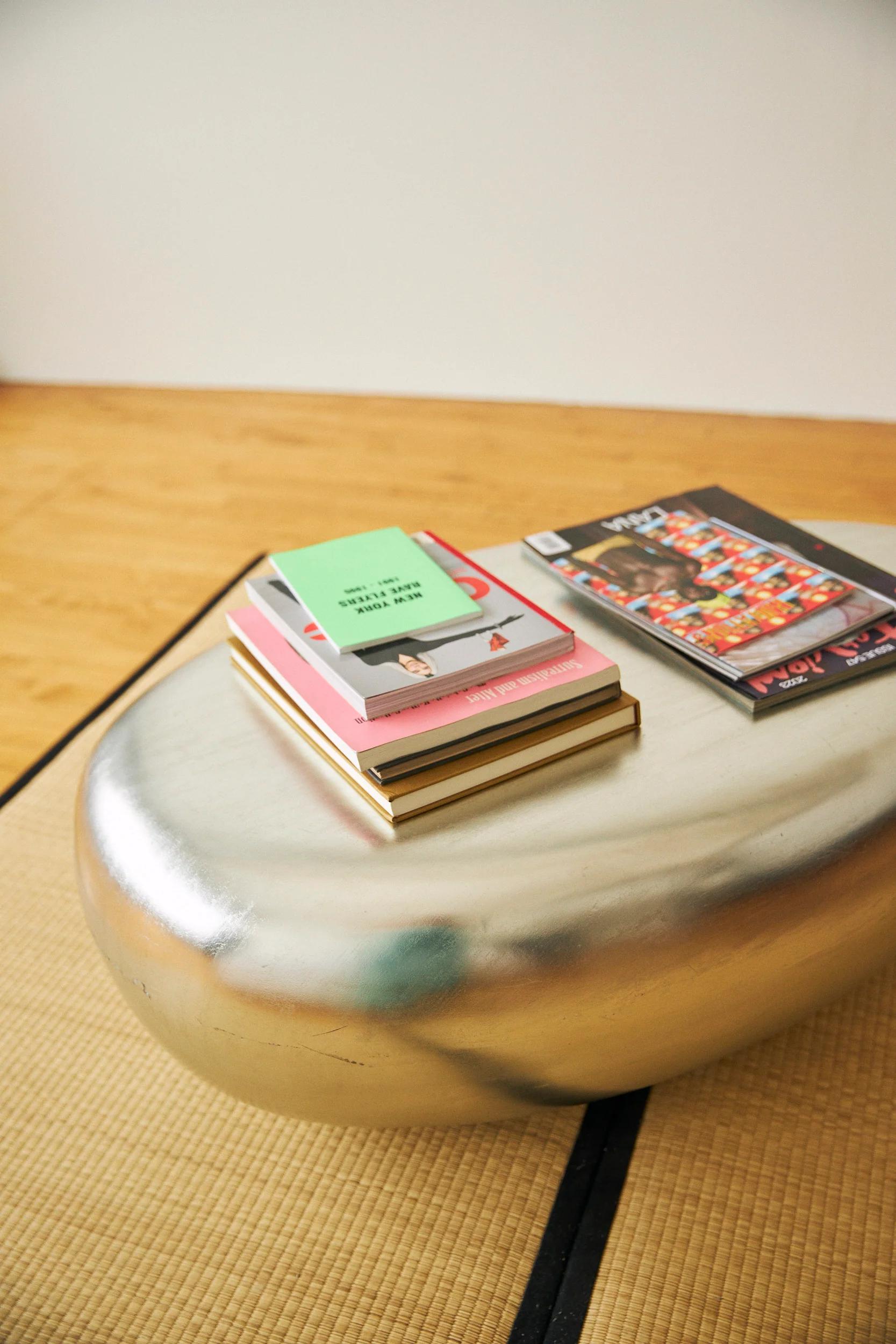 An abstract and minimalist coffee table with a metallic surface on top of a woven rug. Stacks of books and magazines. Features a wooden floor and a blank white wall.