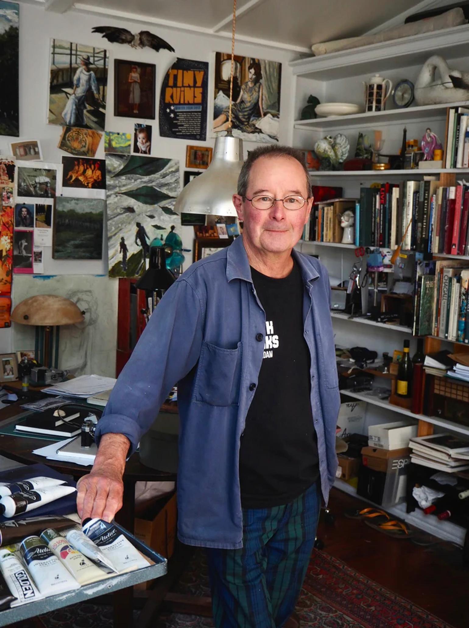 Bespectacled Thomson in casual attire leans on supply-covered table. Bookshelves and artworks line walls of his cluttered Devonport studio.