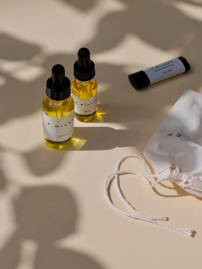 Flatlay of two yellow dropper bottles, lip balm, and white bag. Beige surface with leaf shadows. Skincare or beauty product display.