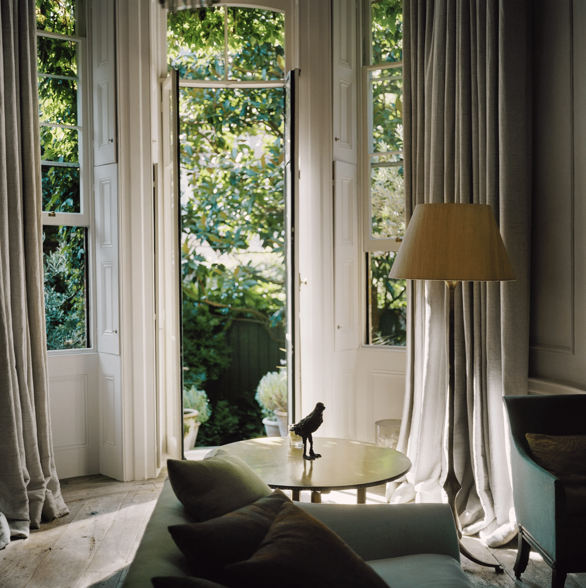 Sunlit room with French doors to garden. Curtains, round table with bird statue, sofa with cushions.