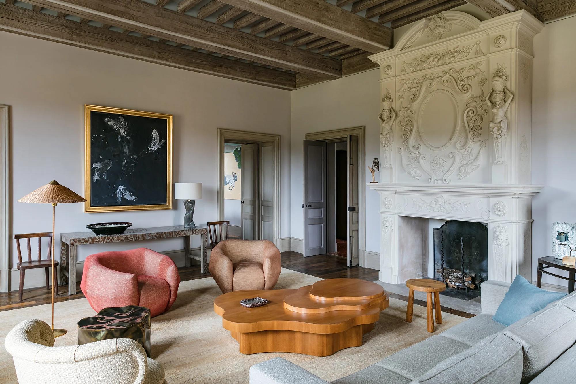 A stylish living room with Provençal charm, featuring a carved fireplace, mixed furniture, and exposed ceiling beams. Modern art adds contrast.