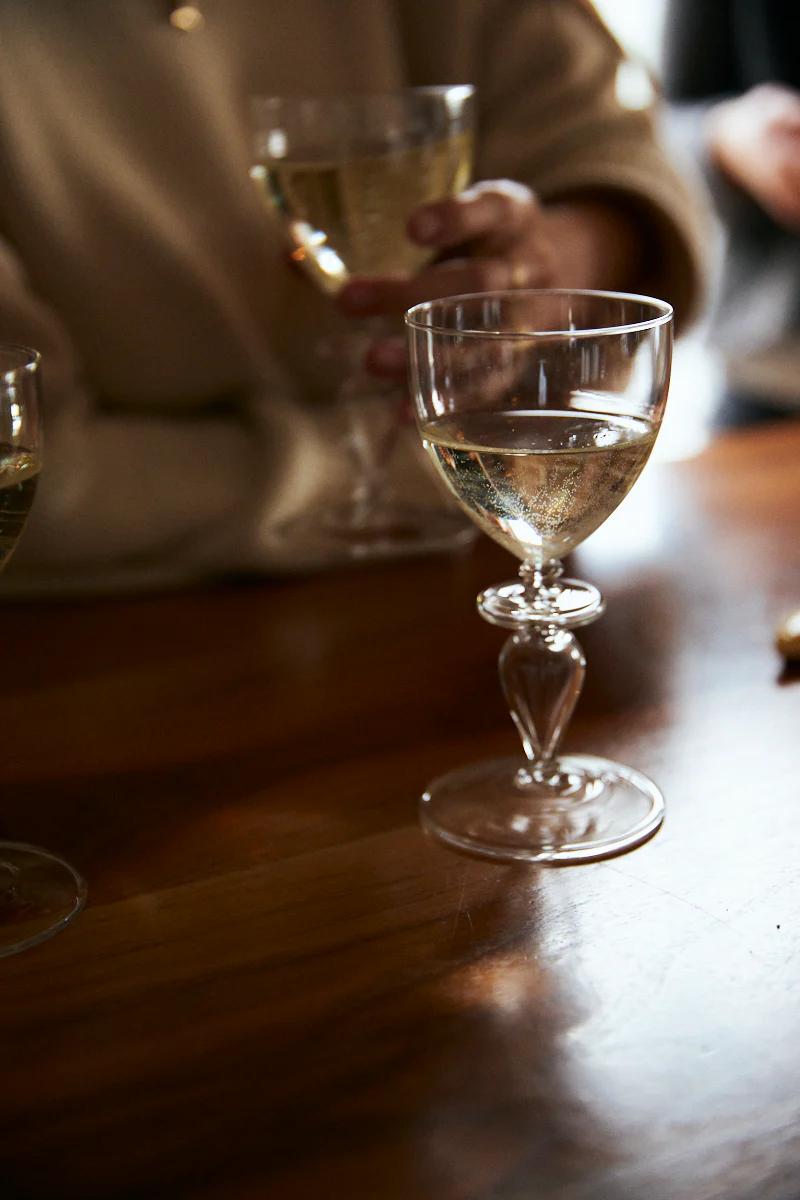 Person holding wine glass, another on table. Jar of mostarda beside glasses in casual indoor setting.