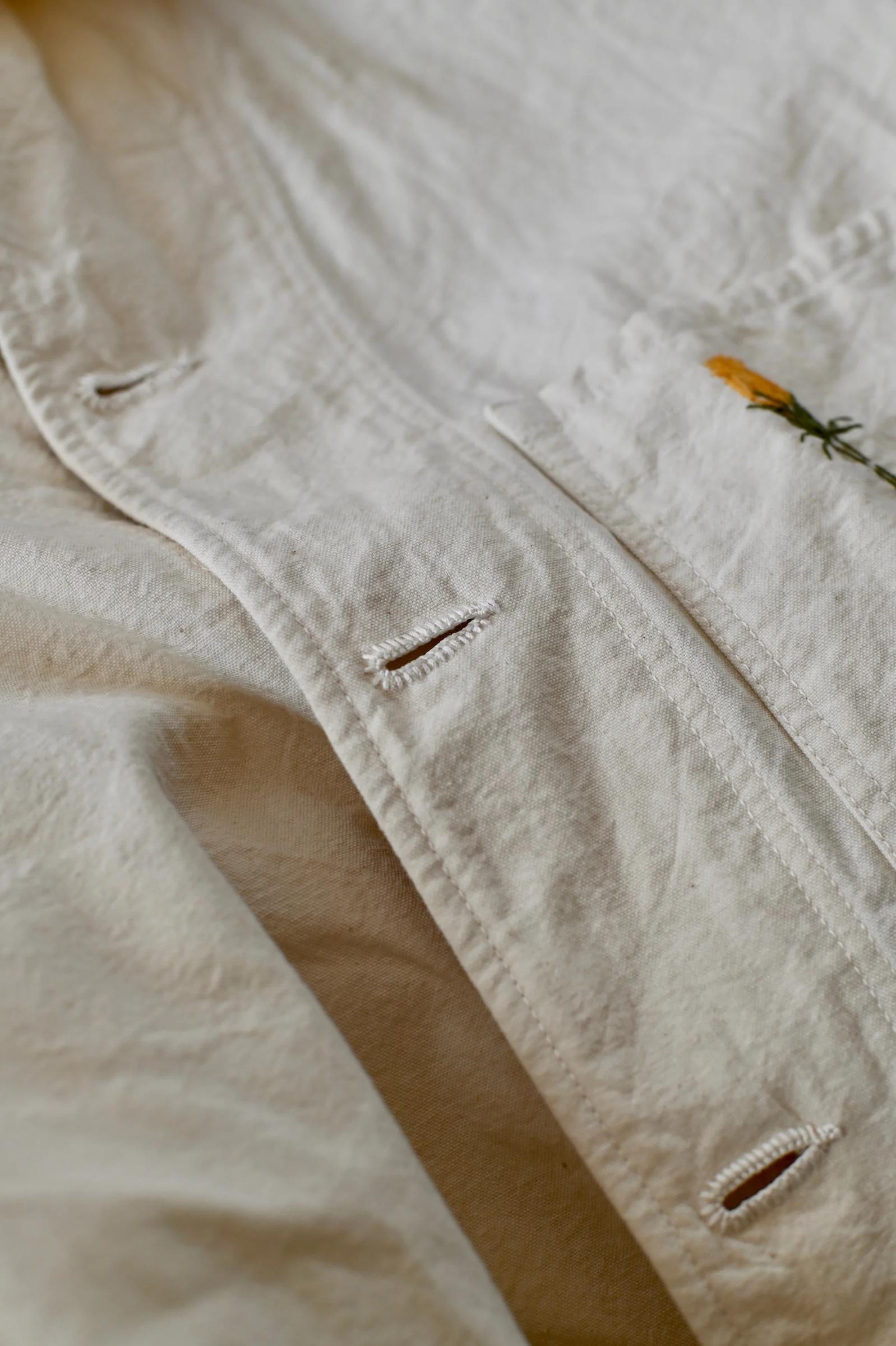 Close-up of beige shirt placket with buttonholes. Small yellow flower embroidered on upper right. Shows Oliver Church's attention to detail.