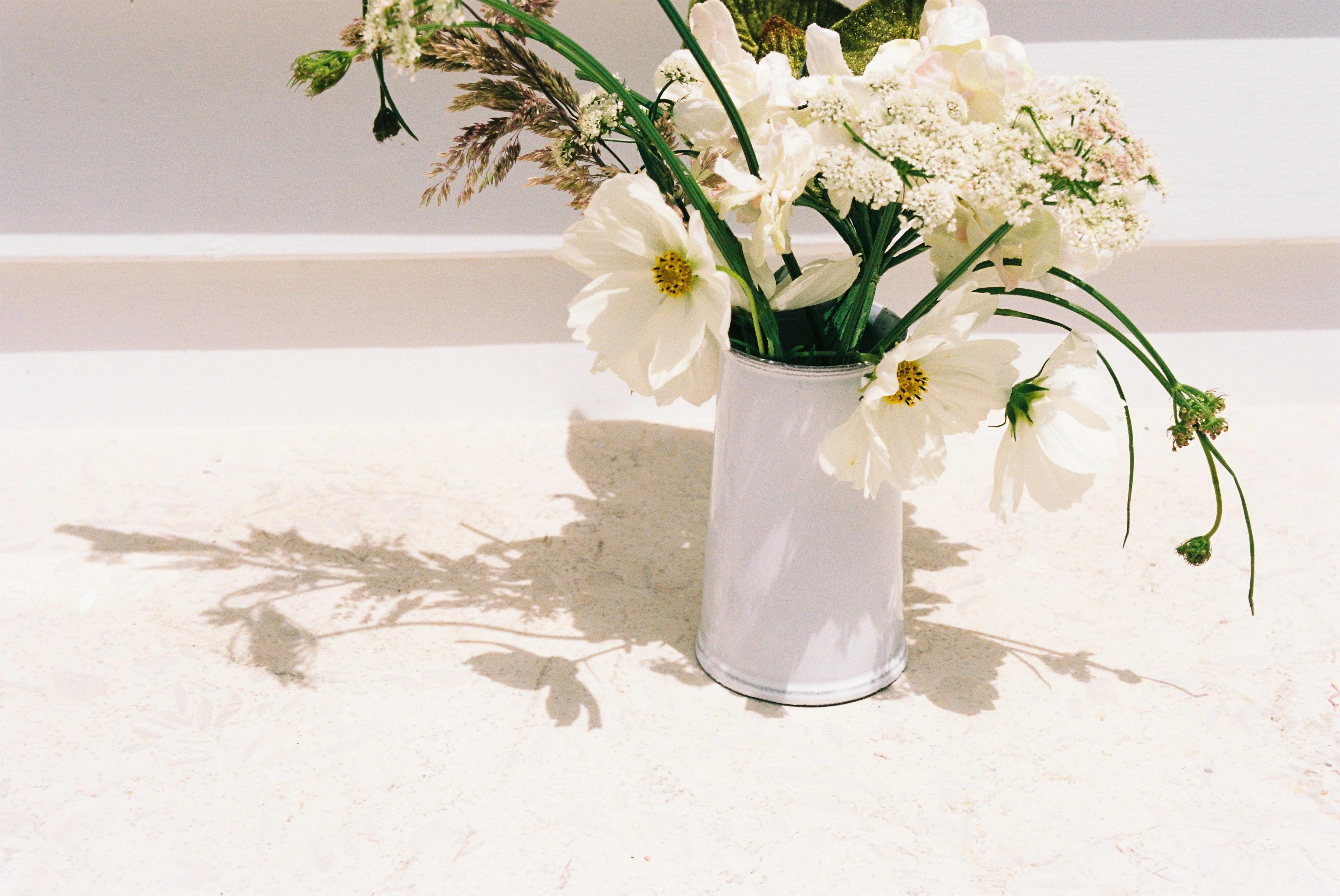 A white vase filled with white flowers and greenery casting soft shadows on a light surface, creating a simple, natural arrangement for festive celebrations.