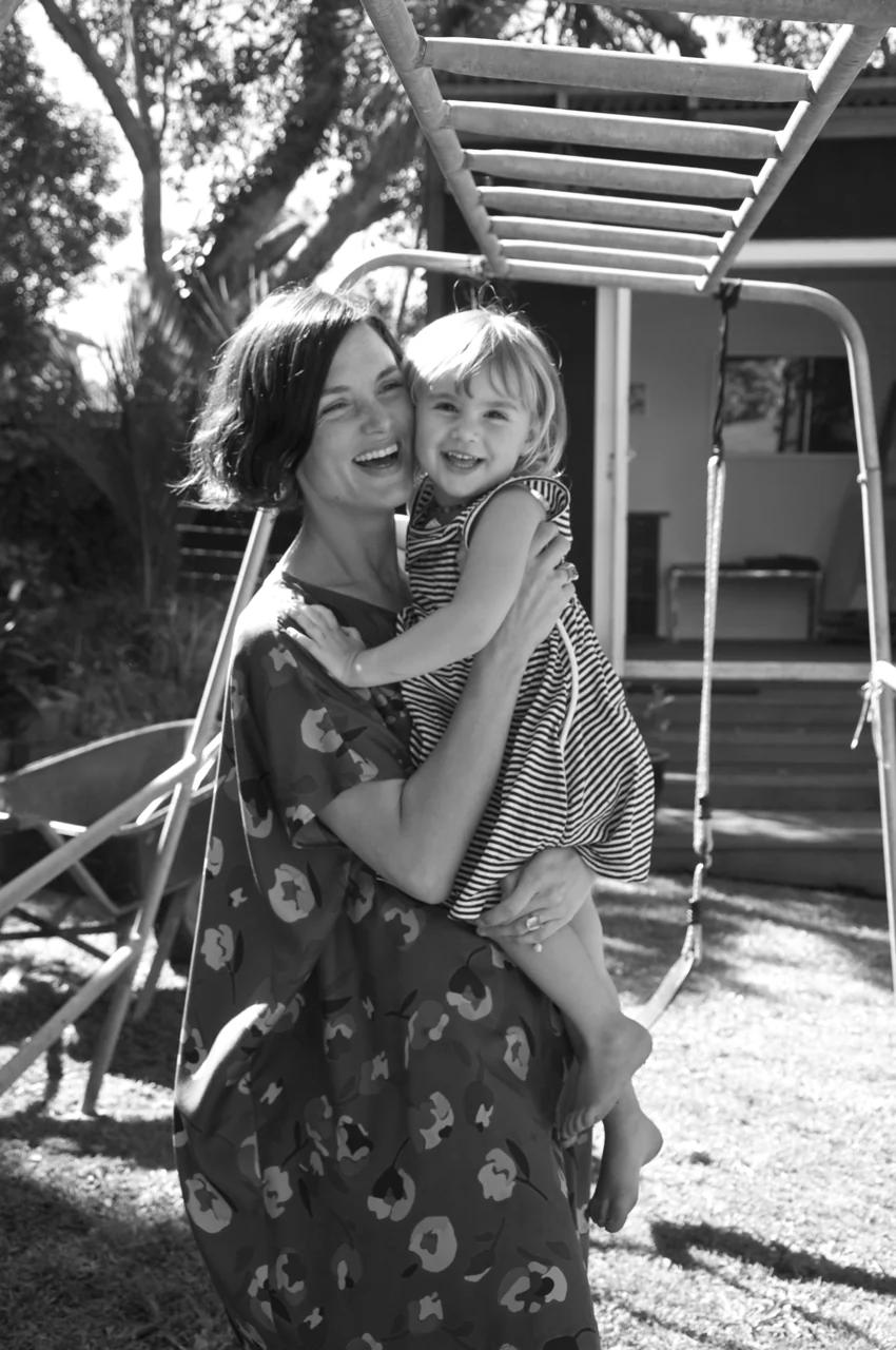 Woman in floral dress holds smiling toddler in striped outfit. Standing by playground equipment in sunlit backyard, creating a joyful family scene.