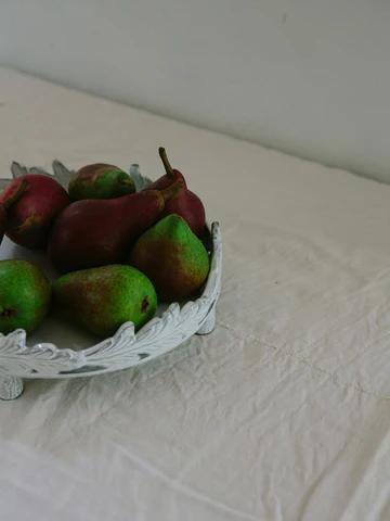 White Astier de Villatte dish on tablecloth holds ripe pears with green and red hues. Simple, rustic presentation against plain background highlights the fruit.