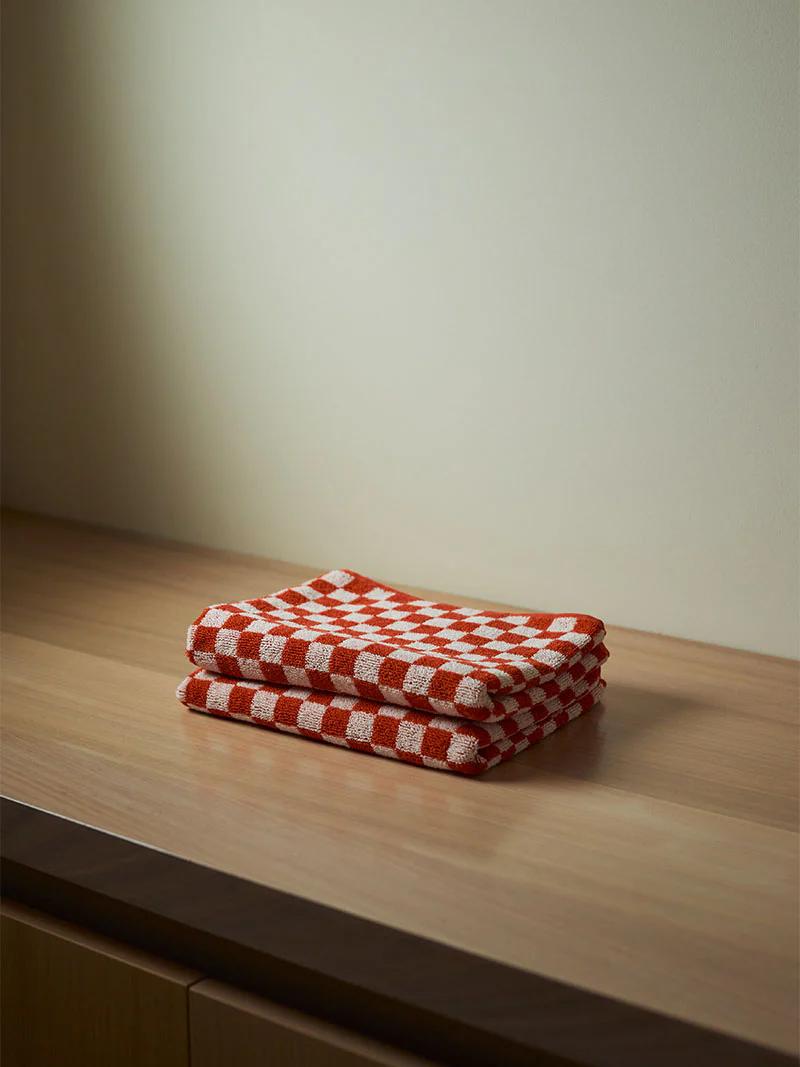 A stack of red and white checkered dish towels neatly folded on a light wooden countertop, set against a plain, light-coloured wall.