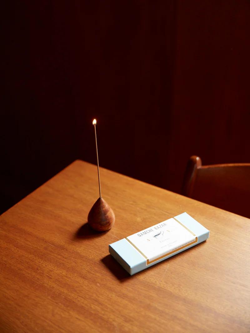 Lit incense stick in wooden holder. Rectangular incense box nearby. Wooden chair partially visible.