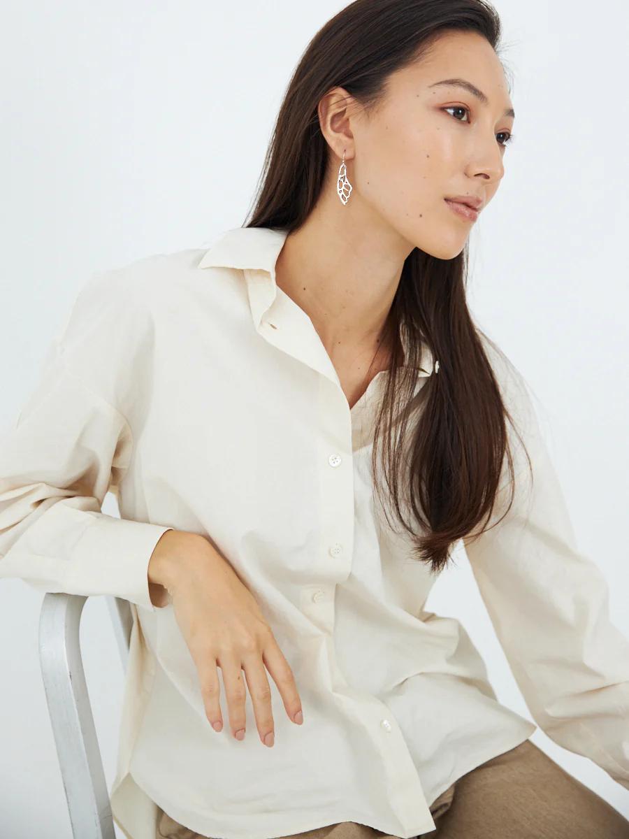 Person with long dark hair in cream shirt wears leaf-shaped earrings. Looking thoughtfully to side. White background.