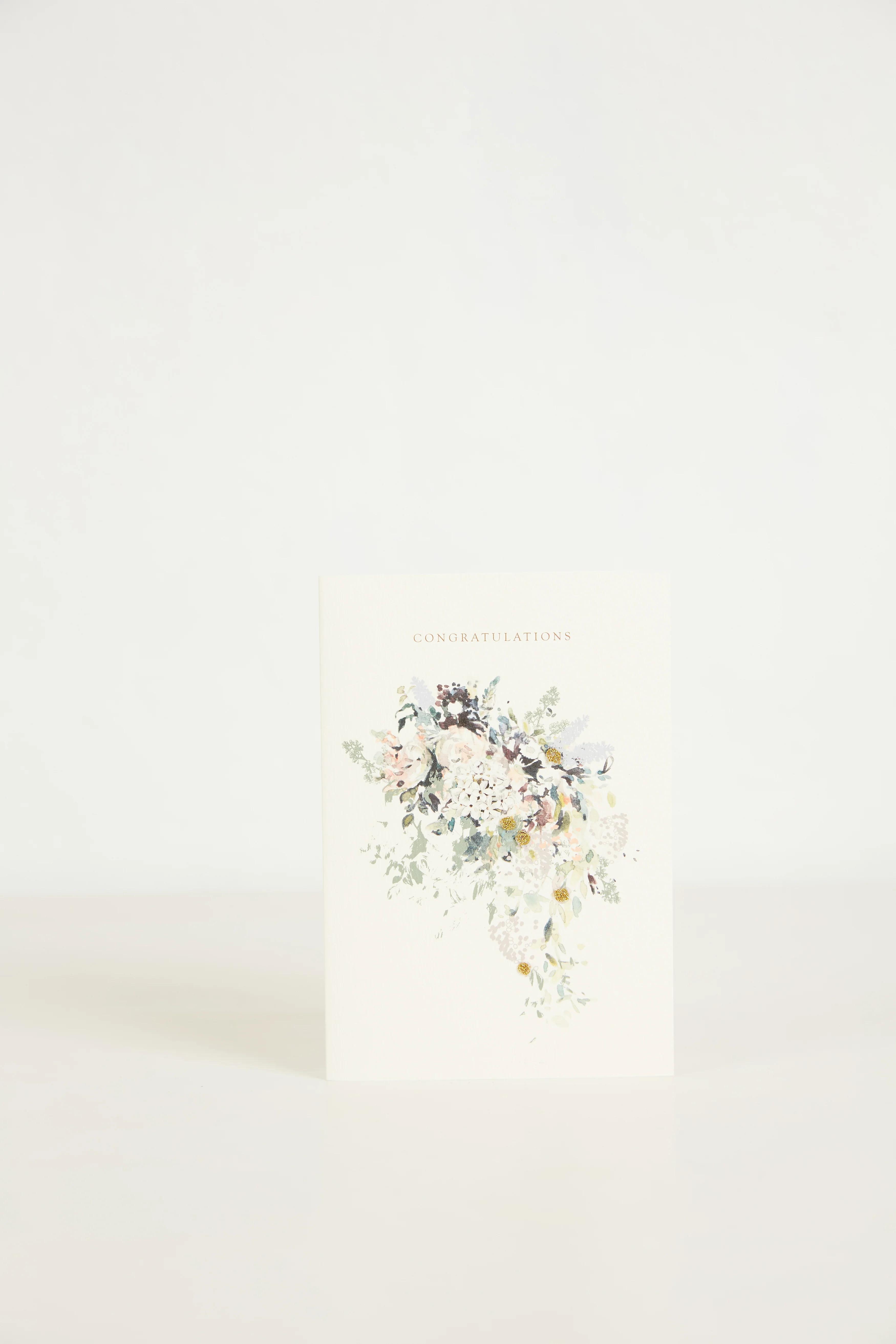 A greeting card with a floral bouquet in pastel colours stands upright against a plain white surface. Above the bouquet is text reading "Congratulations".